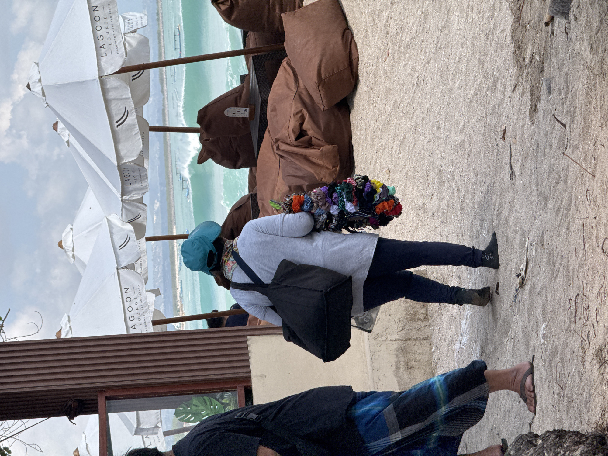 In Kuta, Indonesia, on a beach, a person wearing a blue headscarf and a light gray long-sleeved shirt walks away from the camera. They are carrying a large black tote bag on their left shoulder and a bunch of colorful hair ties in their right hand. Their legs are clad in dark pants. To their left, another person, partially visible from behind, wears a black t-shirt and blue and black patterned sarong. In the background, white beach umbrellas with "Lagoon Lounge" branding are set up, and large brown beanbag chairs are arranged on the sand. The ocean, with gentle waves and several small boats, is visible beyond the seating area. The sky is overcast.