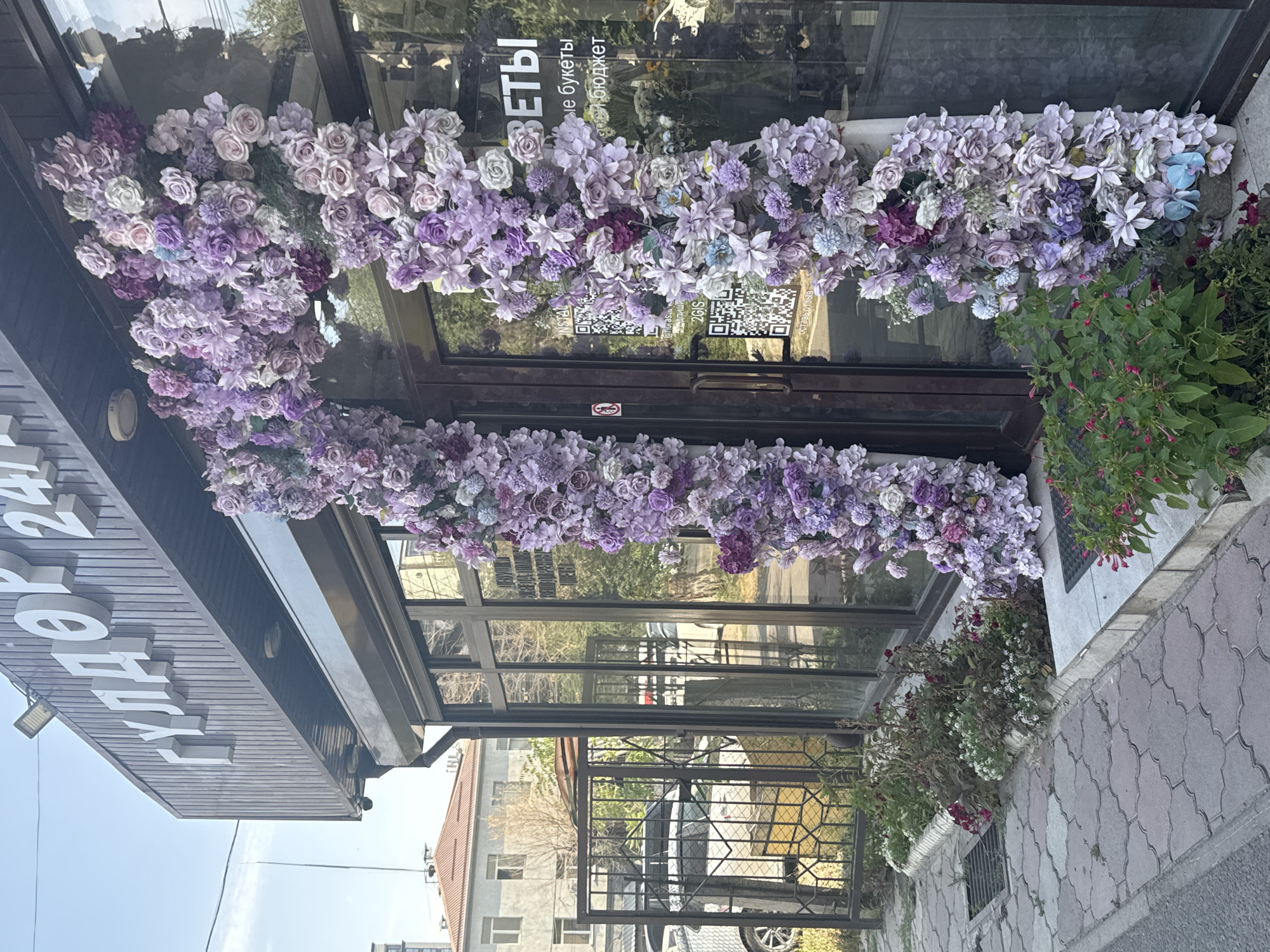The image shows the entrance to a flower shop in Bishkek, Kyrgyzstan. A large archway made of purple and white artificial flowers is draped over the entrance. The shop's name, "ГУЛДОР 24" (GULDER 24), is visible in large white letters on the facade above the entrance. The shop has large glass windows and a dark brown door. QR codes are displayed on the glass. Outside the shop, there is a sidewalk paved with interlocking stone tiles, a metal fence, some plants, and parked cars in the background. The sky is visible and appears to be daytime.