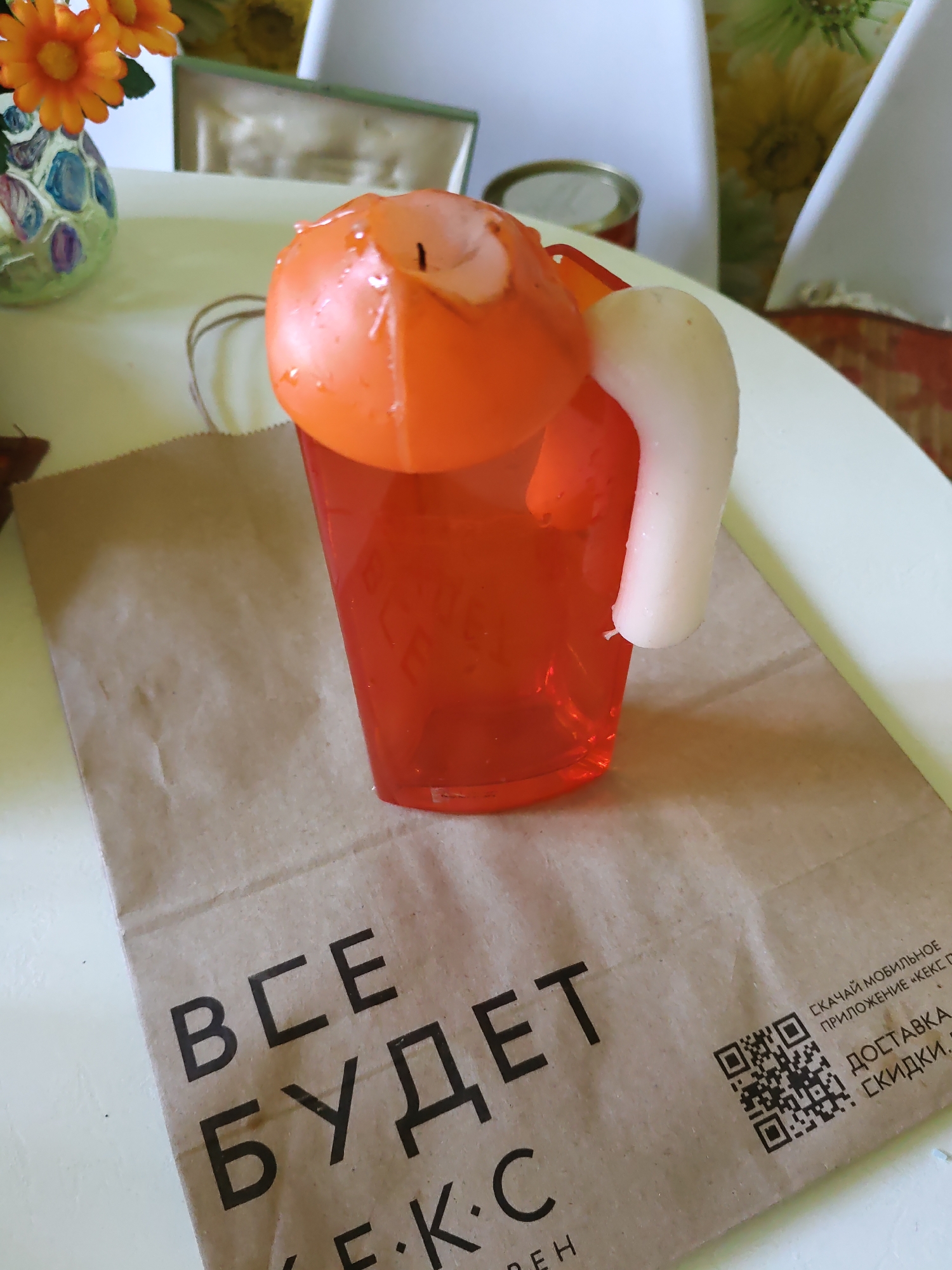 A close-up, slightly overhead shot focuses on an orange candle shaped like a mug of beer, complete with a white foam top, resting on a brown paper bag. The bag has black Cyrillic text printed on it, which appears to be an advertisement for a mobile application and delivery service. To the left of the candle is a colorful vase with orange artificial flowers. The background is blurred, but suggests an indoor setting with hints of furniture and sunlight. The lighting is bright, suggesting daytime. The scene is devoid of people, animals, or any discernible signs of recent activity. The overall impression is of a staged product photograph or a simple still life.