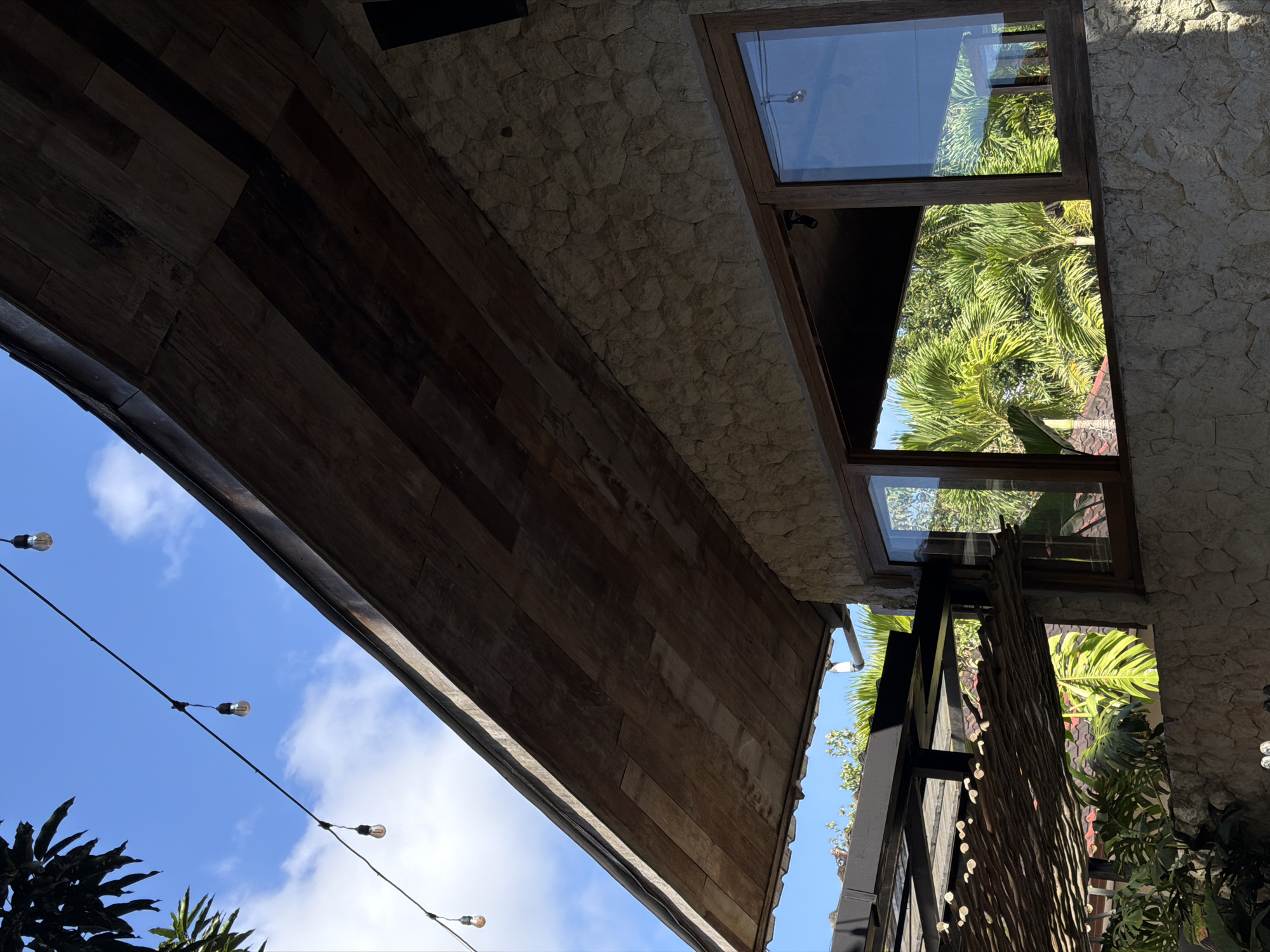 This is a photograph taken outdoors in Kuta Selatan, Indonesia. The image captures a view of a building with a stone facade and a wooden overhang. The overhang is lined with wooden planks, and string lights with exposed bulbs hang from a wire stretched across the sky. Through the windows of the building, lush green palm trees are visible, suggesting a tropical environment. Part of a thatched roof structure and some foliage are also present in the foreground. The sky is a clear blue with some scattered white clouds. The overall impression is of an outdoor seating area or a structure integrated with its natural surroundings.