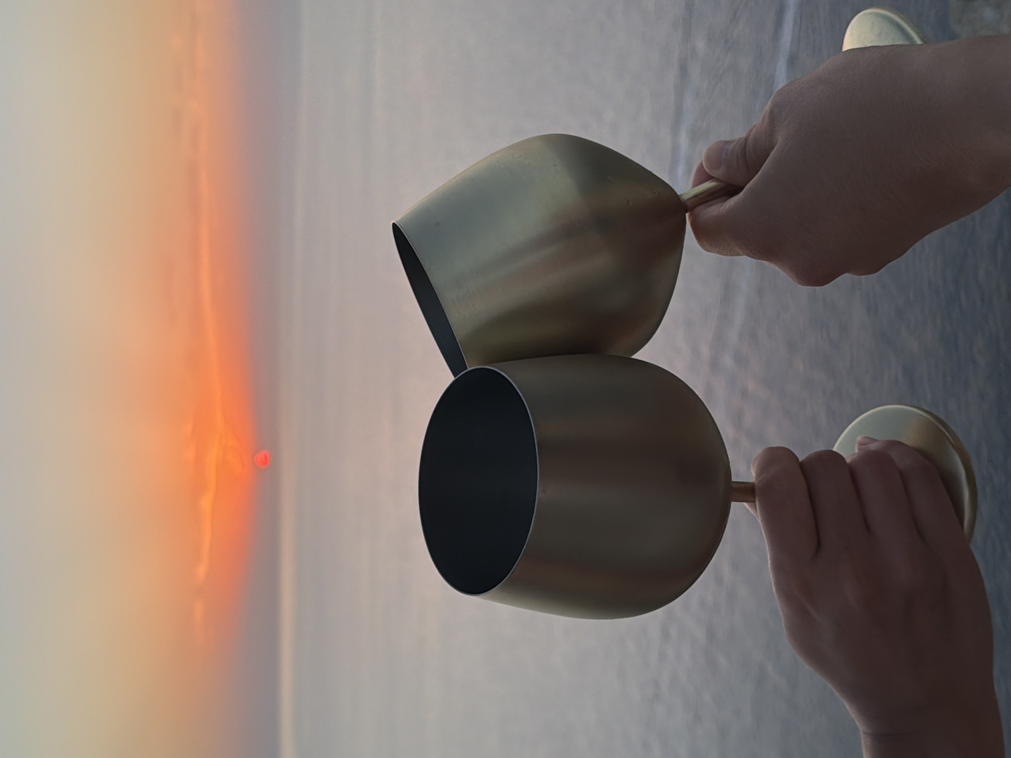 Two metallic goblets are raised in a clinking gesture against a backdrop of the sea and sky at sunset. The sun, a vibrant orange disc, is low on the horizon, casting a warm glow that is reflected in the water. The sky transitions from a pale blue at the top to shades of orange and pink near the horizon. Two human hands are visible, each holding one of the goblets. The goblets appear to be made of a brushed gold-colored metal and have a wide, rounded bowl. The image is a close-up, focusing on the toast with the sunset as a scenic element. The location is identified as Sant Antoni de Portmany, Spain, suggesting a coastal setting, likely a beach or promenade, during the evening.