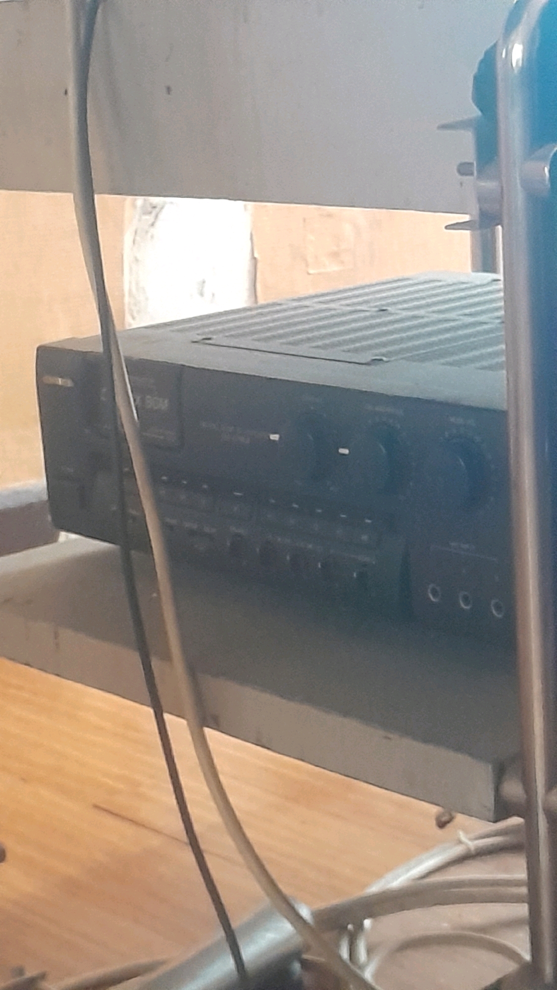 The image shows a close-up, low-angle shot of a black audio receiver unit sitting on a shelf. The receiver has a ribbed top and a front panel with various buttons, knobs, and indicator lights. Text on the front panel is partially visible, including the letters "BGM". Several cables are visible, some draped over the receiver and others lying on the shelf. The shelf appears to be made of metal and is positioned in front of a wall that seems to be constructed of roughly finished material. The lighting is dim, casting shadows on the receiver and the surrounding environment. The floor beneath the shelf is visible and appears to be made of wooden planks. There are no people visible in the image. The image does not contain any specific elements that would allow for the determination of its location within Madiun, Indonesia.