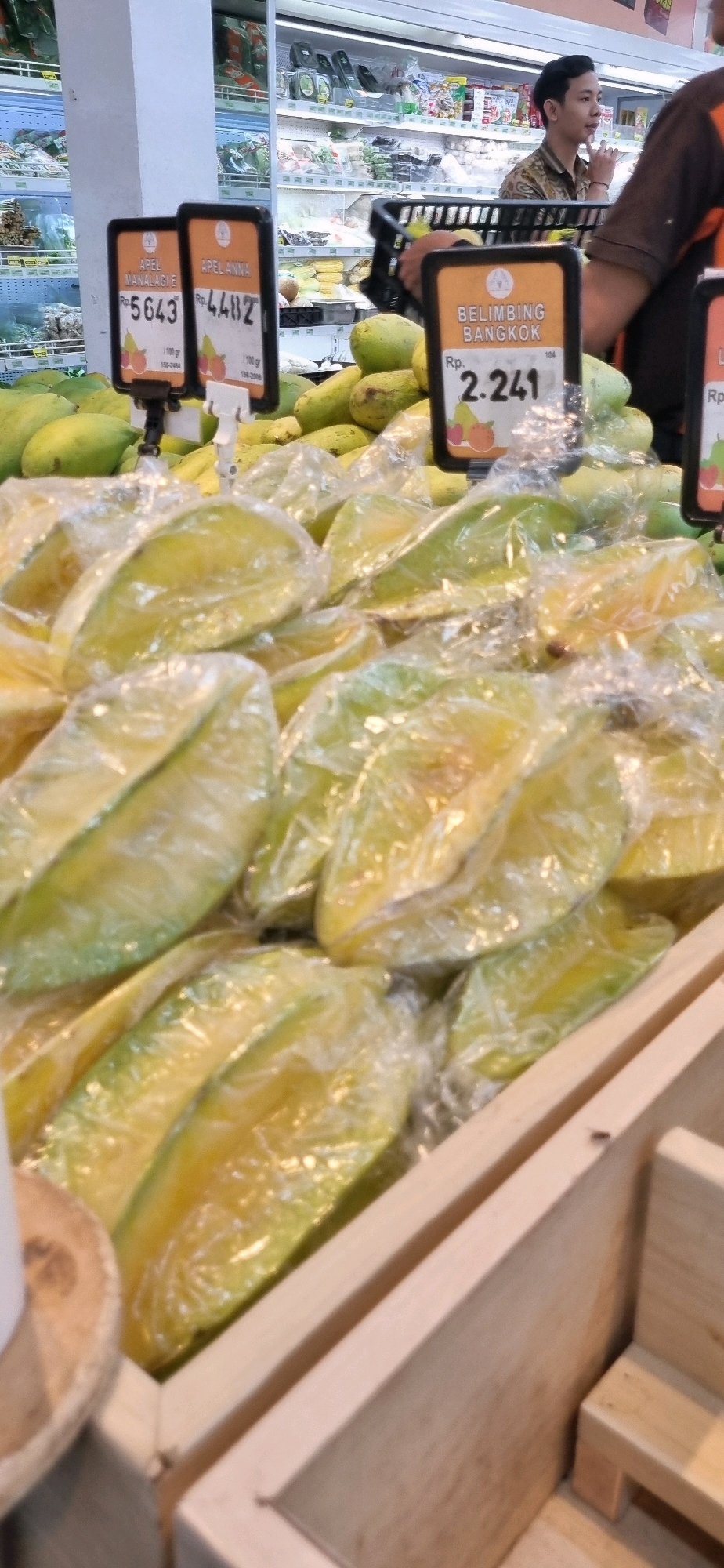 The image depicts a close-up view of star fruits for sale in a market, likely in Denpasar, Indonesia. Several star fruits are visible, individually wrapped in clear plastic. Two price tags are in focus, one for "APEL MANALAGIE" priced at Rp. 5643, and another for "APEL ANNA" priced at Rp. 4482. A third price tag for "BELIMBING BANGKOK" is also partially visible, with a price of Rp. 2.241. In the background, a young man wearing a patterned shirt is looking to the side, and further in the background, shelves stocked with various products can be seen in a brightly lit store. The fruits are displayed in wooden crates.