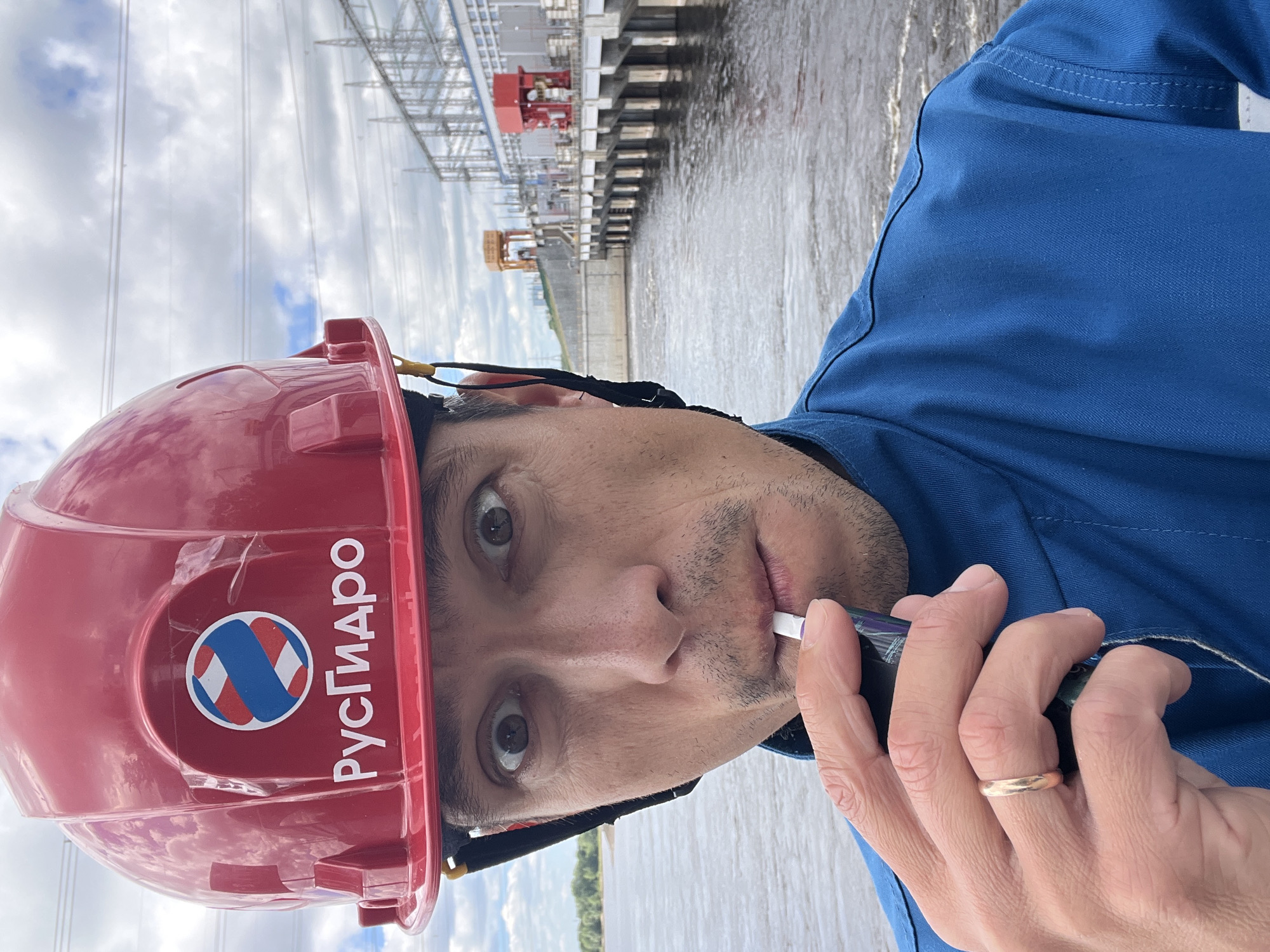 A man wearing a red hard hat with a "RusGidro" logo and a blue uniform is taking a selfie in front of a dam and power station. He is holding a white object to his mouth. The background shows a large body of water, a concrete dam structure, and industrial buildings with tall transmission towers. The sky is cloudy. The location is the Votkinskoye Hydroelectric Station in the city of Chaikovsky, Russia.