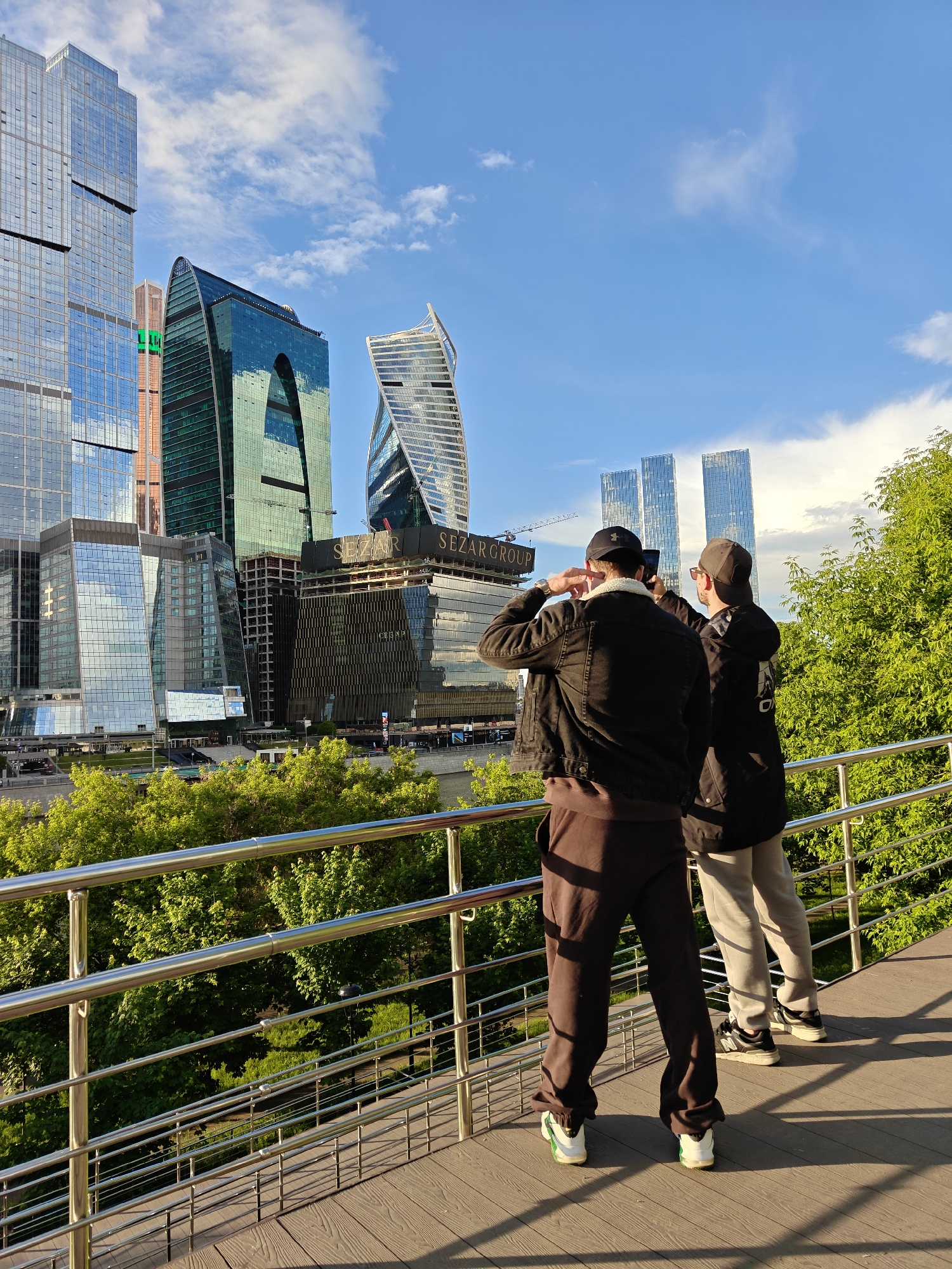 Two people are standing on an elevated walkway, taking photographs of the Moscow International Business Center (Moscow-City) skyline in Moscow, Russia. The walkway has a metal railing. The people are facing away from the camera. The person on the left wears a black baseball cap, a black denim jacket, and brown sweatpants. The person on the right wears a black baseball cap, a black hooded jacket, and gray sweatpants. Green trees fill the middle ground. The Moscow-City skyscrapers are in the background under a blue sky with white clouds. "SEZAR GROUP" is visible on one of the buildings.