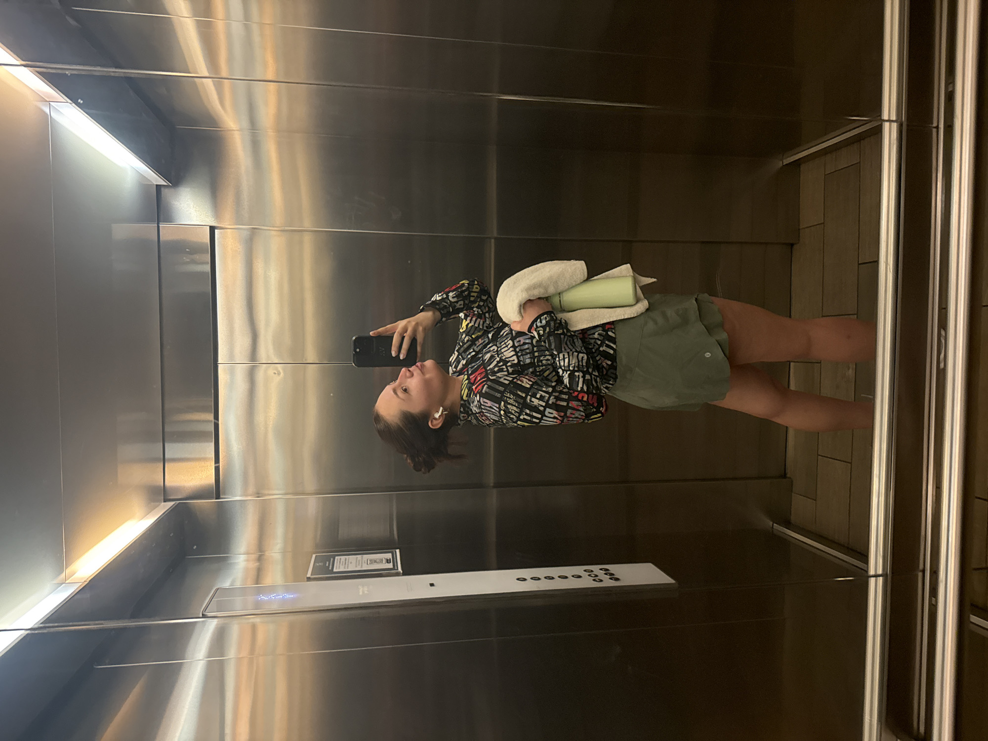 The image shows a person in an elevator, taking a selfie with a smartphone. The person is holding a white towel and a light green water bottle. The elevator has stainless steel walls and a panel of buttons. The elevator floor is tiled. The location is likely in Mexico City, Mexico, based on the available information.