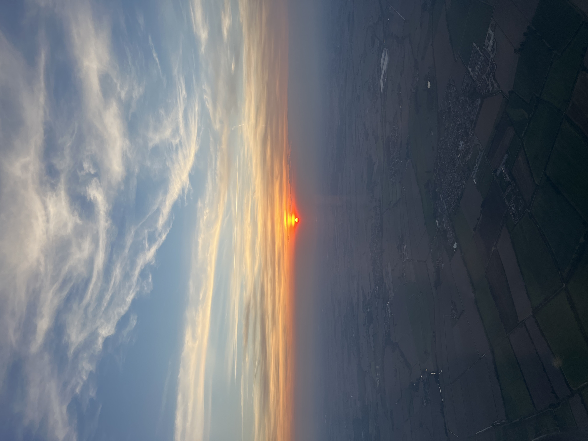 This media file captures a breathtaking aerial view of a landscape during either sunset or sunrise, likely taken from an aircraft. The horizon line runs horizontally across the middle, separating the sky from the terrestrial environment below.
The upper portion of the image features a vast sky, transitioning from a clear light blue at the top to warm, vibrant shades of orange, red, and yellow near the horizon. Delicate, wispy cirrus clouds stretch across the sky, illuminated by the low sun. A faint, linear contrail is also visible in the upper part of the sky. The sun, a brilliant and intense red-orange orb, is prominent on the left side, positioned just above the horizon line, casting a warm glow across the scene and creating a hazy effect near the horizon.
Below the horizon, the landscape consists of an expansive patchwork of agricultural fields. These fields appear in varying geometric shapes and shades of dark green, brown, and grey, indicating cultivated land. Several clusters of buildings, suggesting small villages or rural settlements, are scattered across the terrain. There are also a few small, reflective areas that might be bodies of water or flooded sections. The ground appears darker and somewhat muted due to the low light conditions and atmospheric haze. Based on the provided context, the scene depicts the rural countryside, potentially around Marzano, Italy, at a time of day characterized by dramatic natural light. There is no visible text in the image.