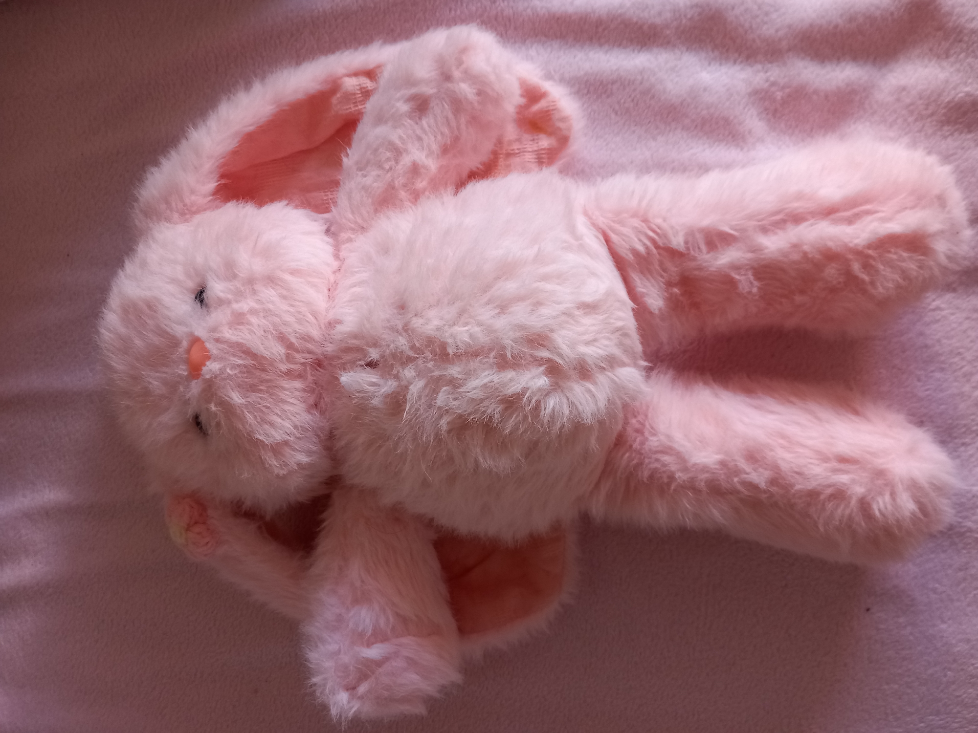 A fluffy pink stuffed bunny is lying on a pink fuzzy blanket. The bunny is lying on its back with its arms spread out to its sides and its legs extended. The bunny has black eyes, a pink nose, and long floppy ears. The background is a pink blanket with a subtle woven texture. The lighting is soft and even, suggesting an indoor environment. The location is Unknown, Unknown.