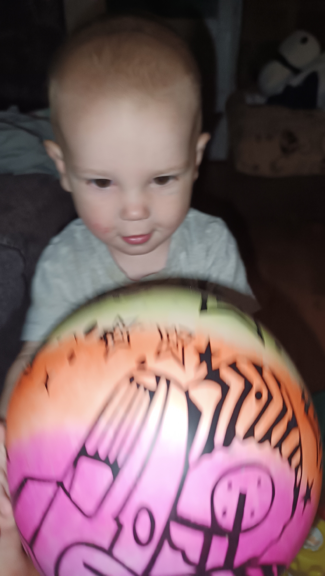 A young child with light brown hair and brown eyes looks down at a colorful ball that is blurred from motion. The child is wearing a light gray t-shirt. The ball is pink, orange, and green with black graffiti-style designs. The background is dark and out of focus, showing what appears to be furniture and possibly a stuffed animal. The image is taken from a close-up perspective, with the ball dominating the foreground. The scene is likely indoors. The image does not contain any information that would allow for the determination of its location in Karsha, Russia.
