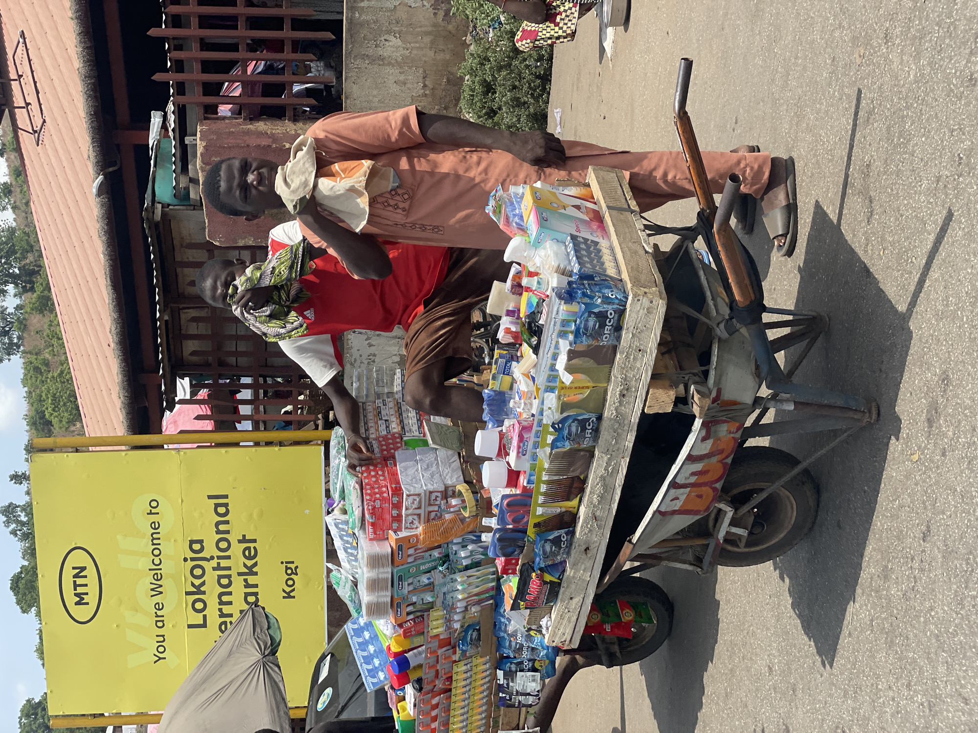 The image shows two men standing next to a cart full of goods in Lokoja, Nigeria. A yellow sign behind them reads "MTN, You are Welcome to Lokoja International Market, Kogi." The cart is stocked with various household and personal care items. One man wears a red shirt and has a scarf around his neck, while the other wears an orange outfit and carries a cloth over his shoulder. The cart is on a paved street.