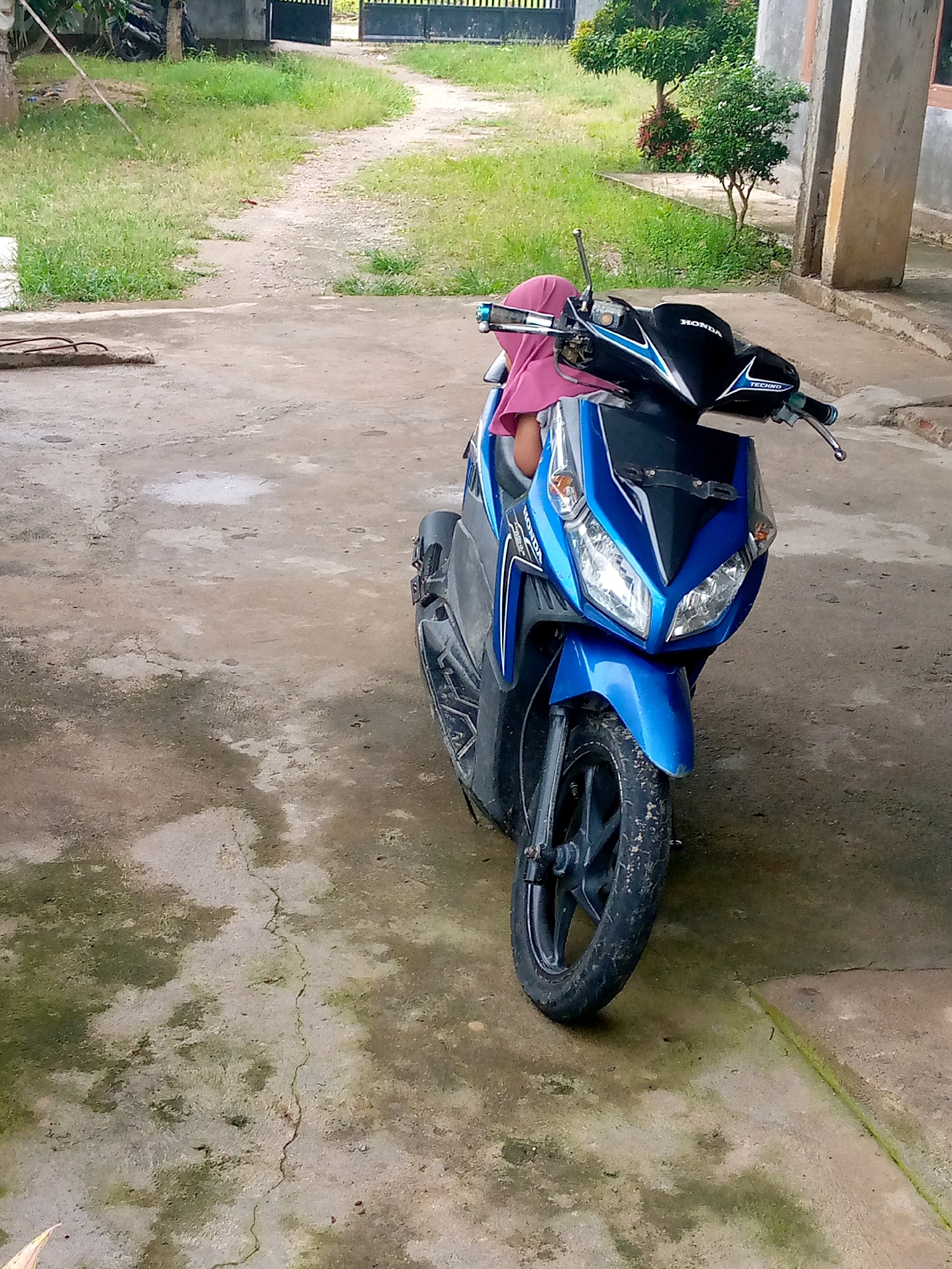 A blue and black Honda scooter is parked on a concrete surface. A person wearing a pink headscarf is seated on the scooter, with their left arm resting on the handlebars. The scooter's headlights and front fender are visible. The background shows a grassy area with a dirt path leading to a gate. There are trees and a building in the distance. The concrete surface is stained and cracked in several places, with some moss growth. The lighting suggests an outdoor scene during daylight hours.