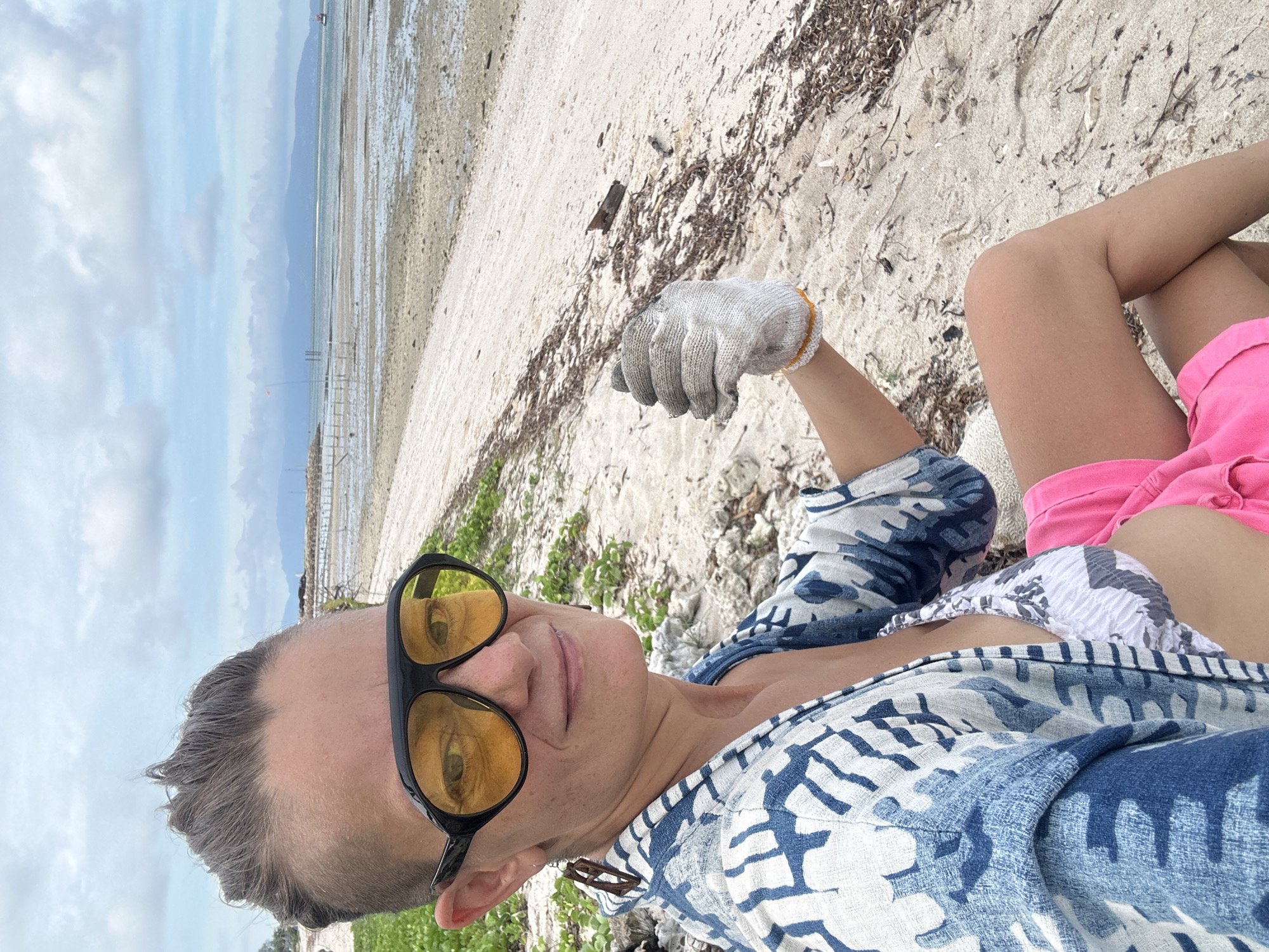 A woman with a short, shaved haircut and yellow aviator sunglasses is seated on a sandy beach, looking towards the camera with a slight smile. She is wearing a blue and white patterned robe over a bikini top and bright pink shorts. Her right hand, covered by a gardening glove, is raised and positioned in front of her. The background shows a wide, tranquil beach with scattered debris, calm ocean water, and distant islands under a cloudy sky. The scene suggests an outdoor activity, possibly beach cleanup or relaxed beachwear.
