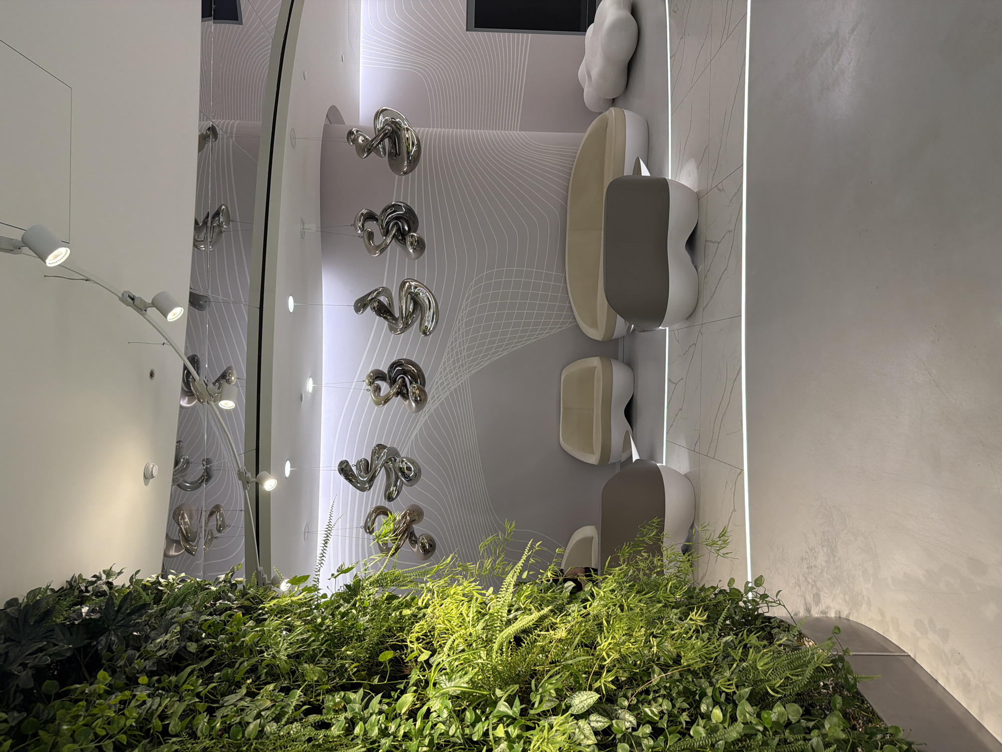 A slightly rotated interior view shows a vertical garden at the bottom, and abstract sculptures and design features hanging from the wall. The color palette is primarily light and muted.
The lower portion of the image is dominated by a dense vertical garden of various green plants and ferns.
Above the garden are several large, chrome abstract sculptures suspended from the wall, appearing to be floating in the air. The wall behind the sculptures features a stylized, wave-like pattern.
To the right of the sculptures are a series of chairs, one of which is a long bench, in a minimalist design. The chairs are composed of light colors with soft, curved forms. Near the top of the image is a decorative, cloud-shaped object.
The walls are a combination of light grey and white, with a vertical strip of LED lighting illuminating the seam between two panels.
There are no people visible in the image.
The location of this image is Moscow, Russia.