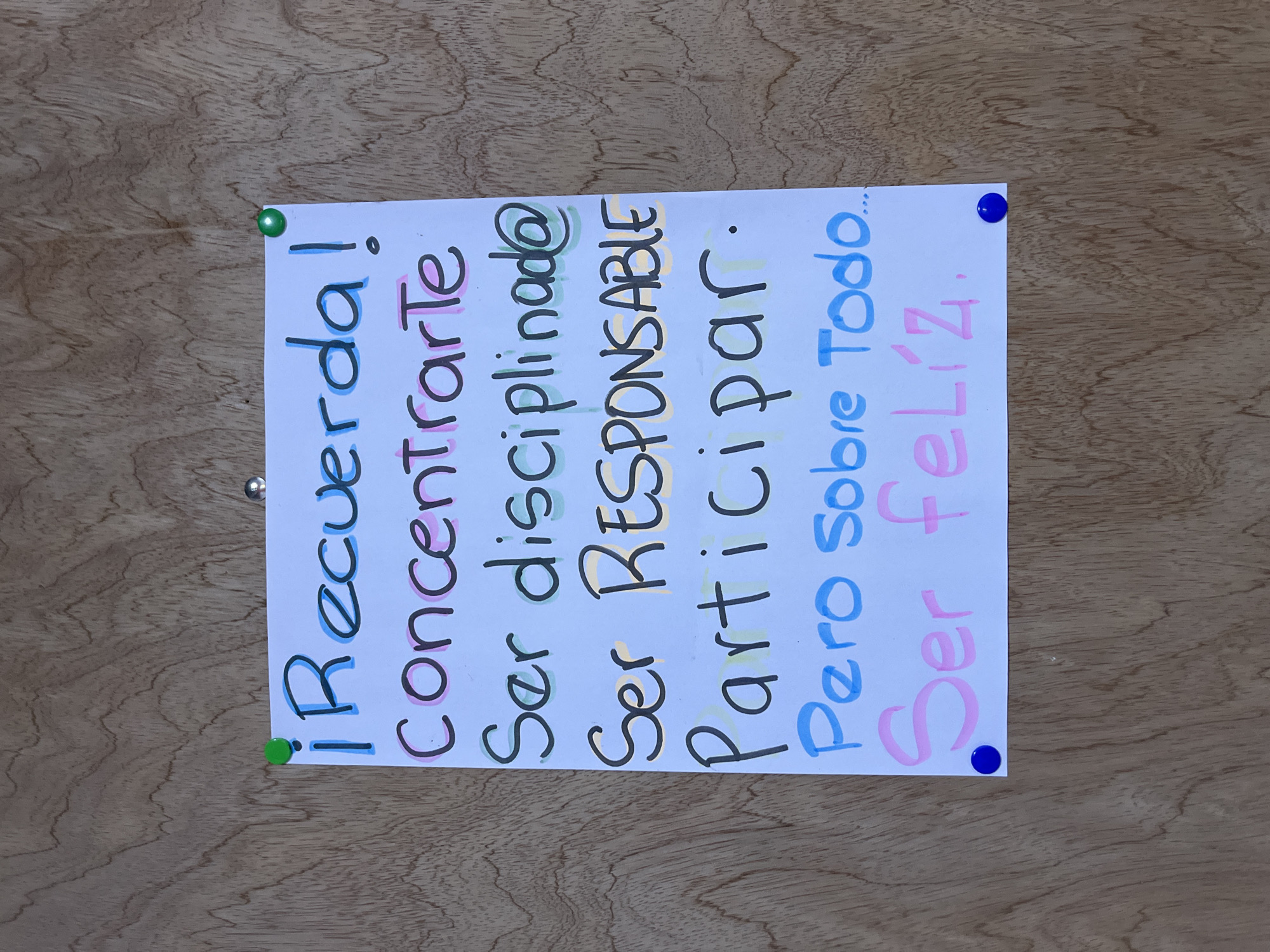 A white piece of paper with handwritten Spanish text is pinned to a wooden surface, likely a bulletin board or wall. The text, written in multi-colored marker, lists reminders: "¡Recuerda!" (Remember!), "Concentrarte" (Concentrate), "Ser disciplinado@" (Be disciplined), "Ser RESPONSABLE" (Be RESPONSIBLE), "Participar." (Participate.), "Pero Sobre Todo..." (But Above All...), and "Ser feliz." (Be happy.). The letters have different colored outlines, with the main text in black. The paper is secured by two green pushpins at the top and two blue pushpins at the bottom. The wooden surface has a visible grain pattern. The image appears to be taken in an outdoor setting, possibly a school or community center, given the subject matter of the poster. This is a static scene with no people present.