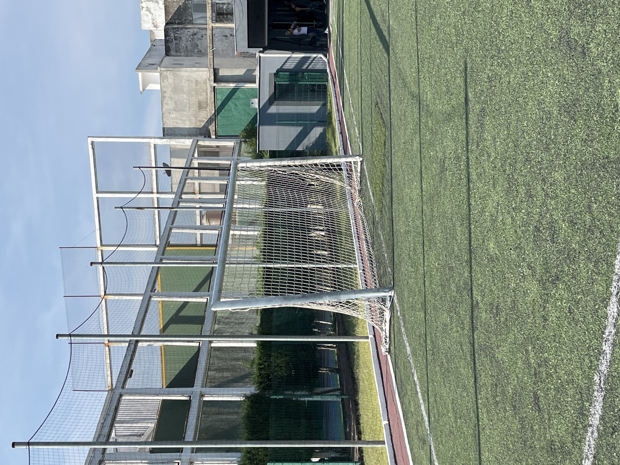 The image depicts an artificial turf soccer field under a clear blue sky on a bright, sunny day. A white metal soccer goal with a net stands prominently in the foreground on the left. The green artificial turf field, marked with white lines, stretches towards the right, bordered by a reddish track or pathway. Behind the goal and to its left, a tall metal framework supports extensive netting, likely serving as a protective barrier for the facility.
In the background, beyond the netting, several multi-story concrete buildings with windows and some green paneling or grates are visible. Trees and bushes are also seen behind the fencing, bordering the field. A few individuals are faintly discernible on a balcony or observation deck of one of the buildings, appearing to be observing the field or resting.
No active game or event is taking place on the field. The bright sunlight casts strong shadows, indicating it is likely mid-morning or afternoon. The overall setting appears to be an urban or semi-urban sports complex. There is no visible text in the image.