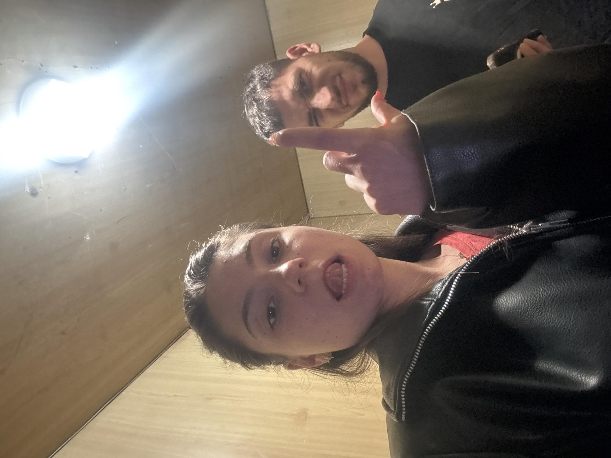 The image is a low-angle selfie taken by a woman indoors, showing a man in the background. The woman is wearing a black leather jacket and a red top. She has dark hair pulled back and is sticking her tongue out. The man in the background has short dark hair and a beard and is wearing a black t-shirt. He is pointing with his index finger. There is a bright light fixture visible on the ceiling. The location appears to be indoors, with wooden paneling on the walls. The city and specific location are unknown.