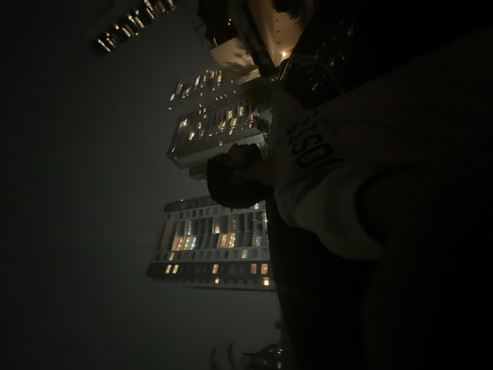 The photo shows a man looking up at two high-rise buildings at night in Huixquilucan de Degollado, Mexico. The buildings are lit with many windows. The man is wearing a light-colored sweatshirt with lettering.