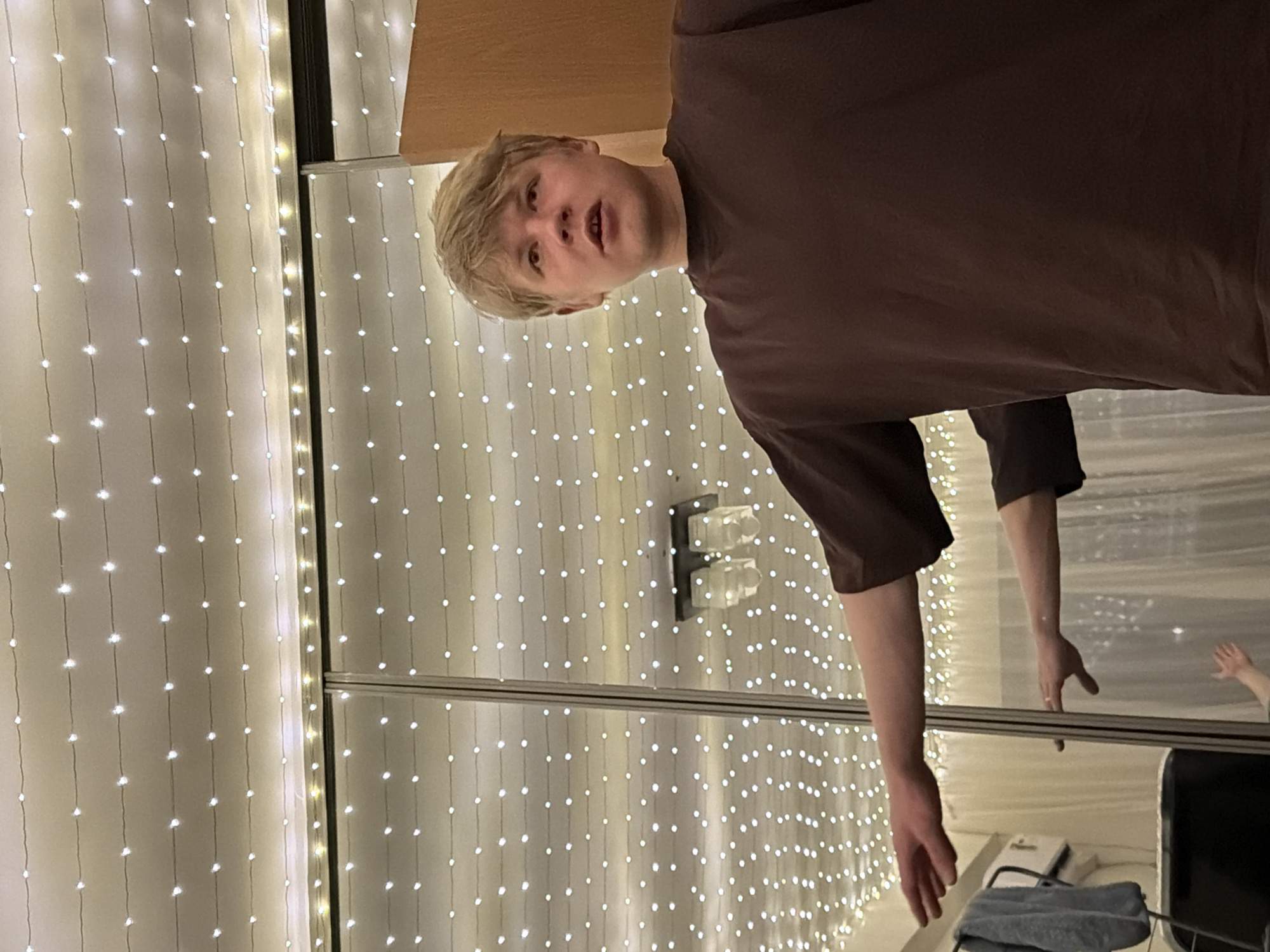 A person with light blonde hair and wearing a brown t-shirt is standing in front of a sliding mirrored wardrobe. The mirrored doors reflect the person, as well as the opposite wall, which is adorned with a curtain of small white lights. On the floor in front of the wardrobe is a desk with a towel or cloth resting on it. The person is the primary subject, with the background composed of the lights, wardrobe, and mirrored reflection. The location of the city of Izhevsk, Russia is not discernable from the visual information provided.