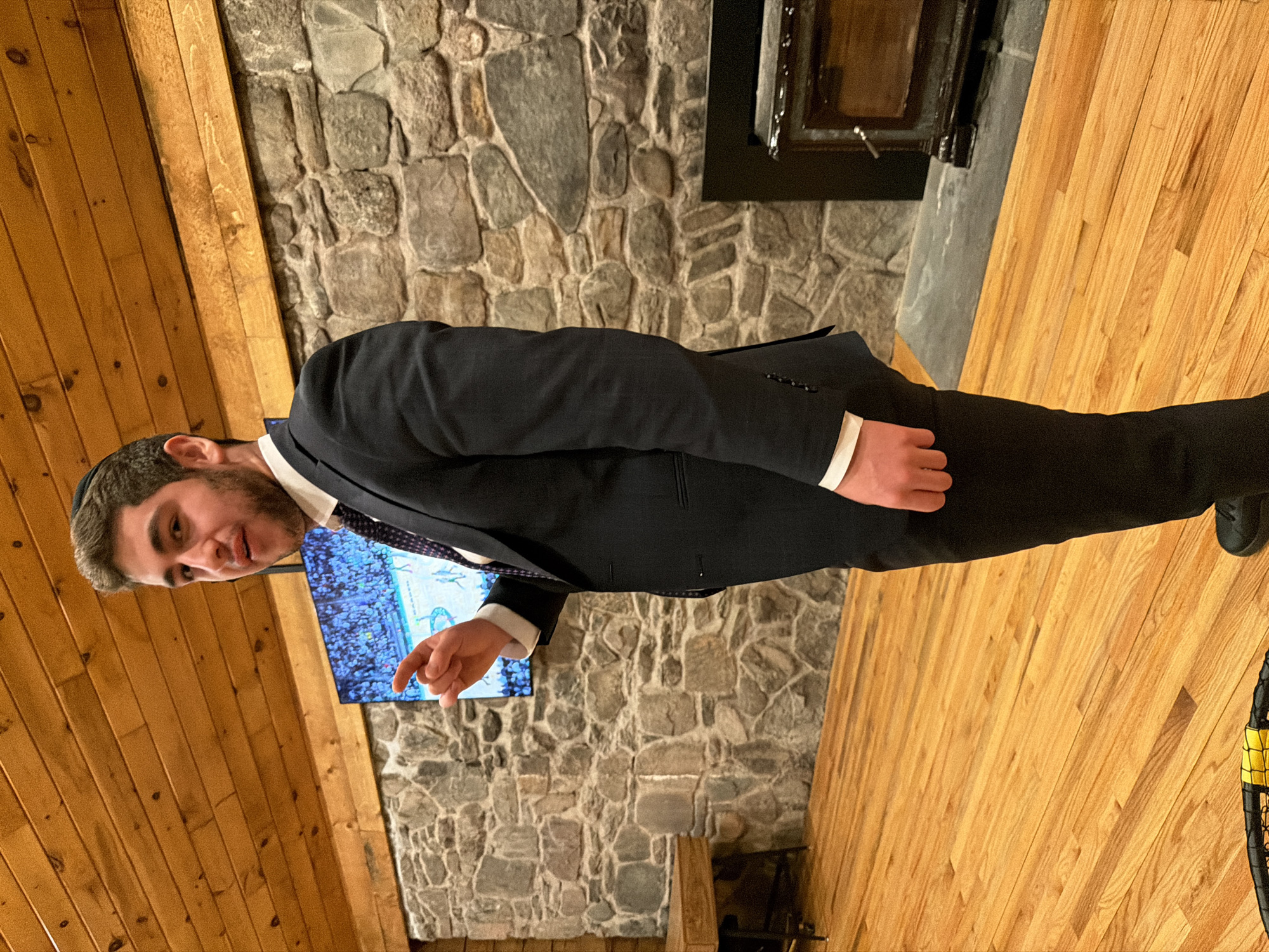 A person in a suit and yarmulke stands in a room with wooden walls and floors, pointing at a TV on a stone wall. There is a fireplace on the stone wall. The location is Rock Hill, United States of America.