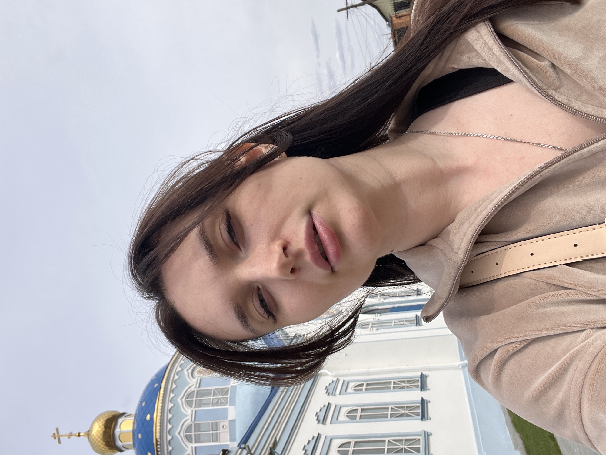 The image shows a woman in Kargopolye, Russia, with a church in the background. The woman has long brown hair and is wearing a light brown jacket and a silver necklace. She is looking slightly downward at the camera. Behind her is a white building with blue accents and a gold dome, which identifies it as an Orthodox church. The sky is overcast. The image is angled, giving the impression that the camera was held at a slight tilt during the shot.