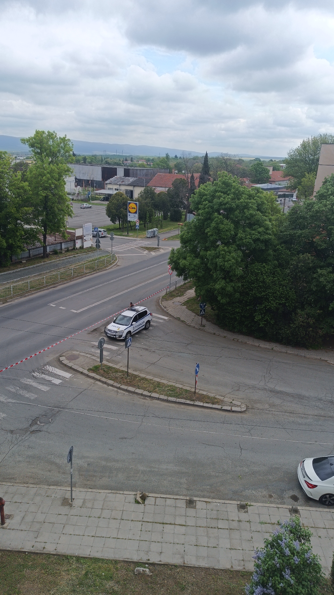 The image shows an intersection in Karnobat, Bulgaria, potentially near the Lidl supermarket. A white police car with "ПОЛИЦИЯ" markings is positioned at the intersection, which is cordoned off with red and white tape. A person can be seen standing next to the car. The environment is urban with buildings, trees, sidewalks, and a visible Lidl store in the background. The sky is cloudy. A white car is parked to the right on the bottom of the photo.