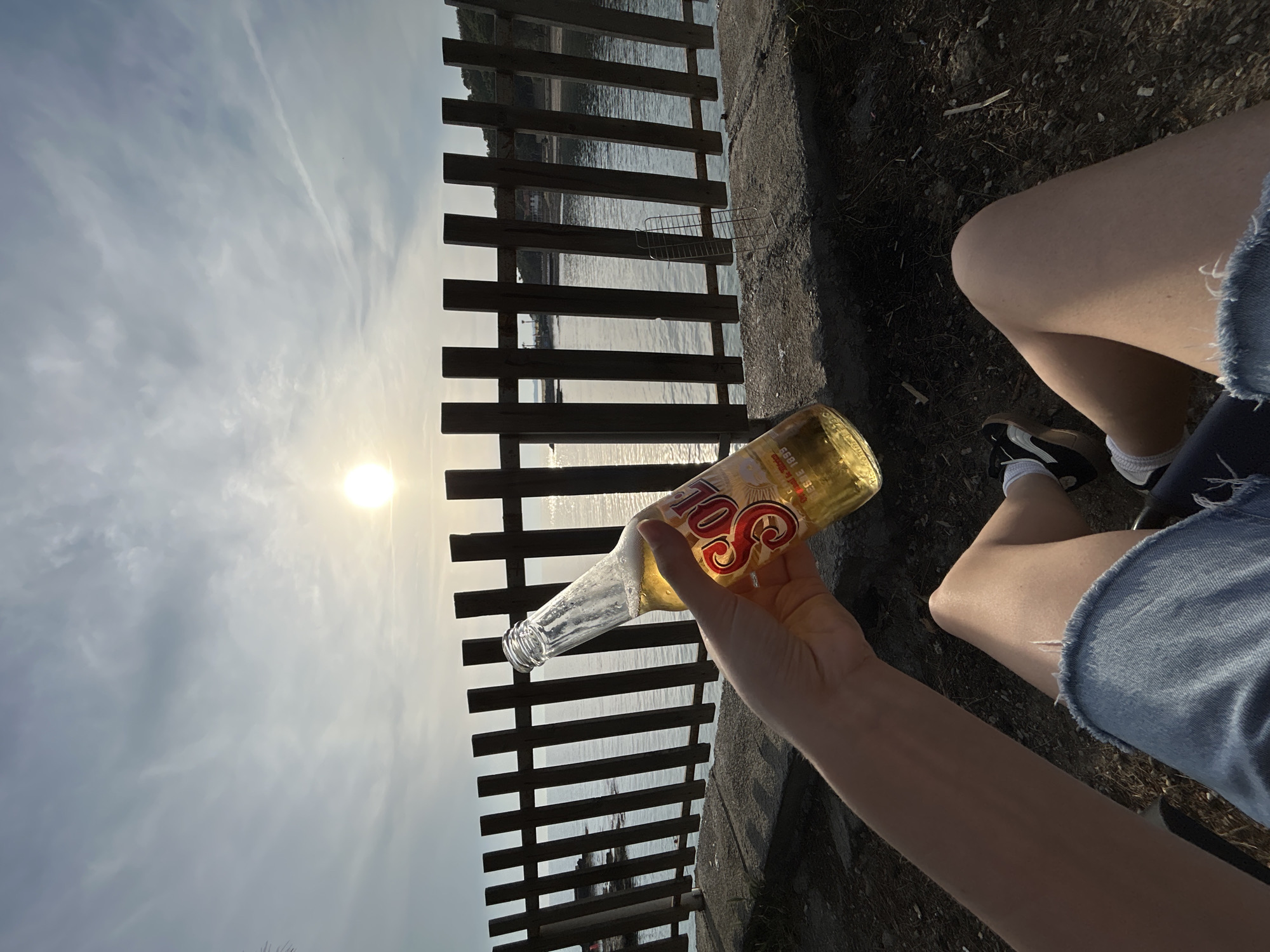 A person is sitting outdoors, holding a nearly empty bottle of Sol beer up to the sun. The background includes a wooden fence and a body of water under a cloudy sky, with the sun visible. The person is wearing a denim short and black and white sneakers. Based on the details of the landscape and sky, this image does not appear to be from Alanya, Turkey.