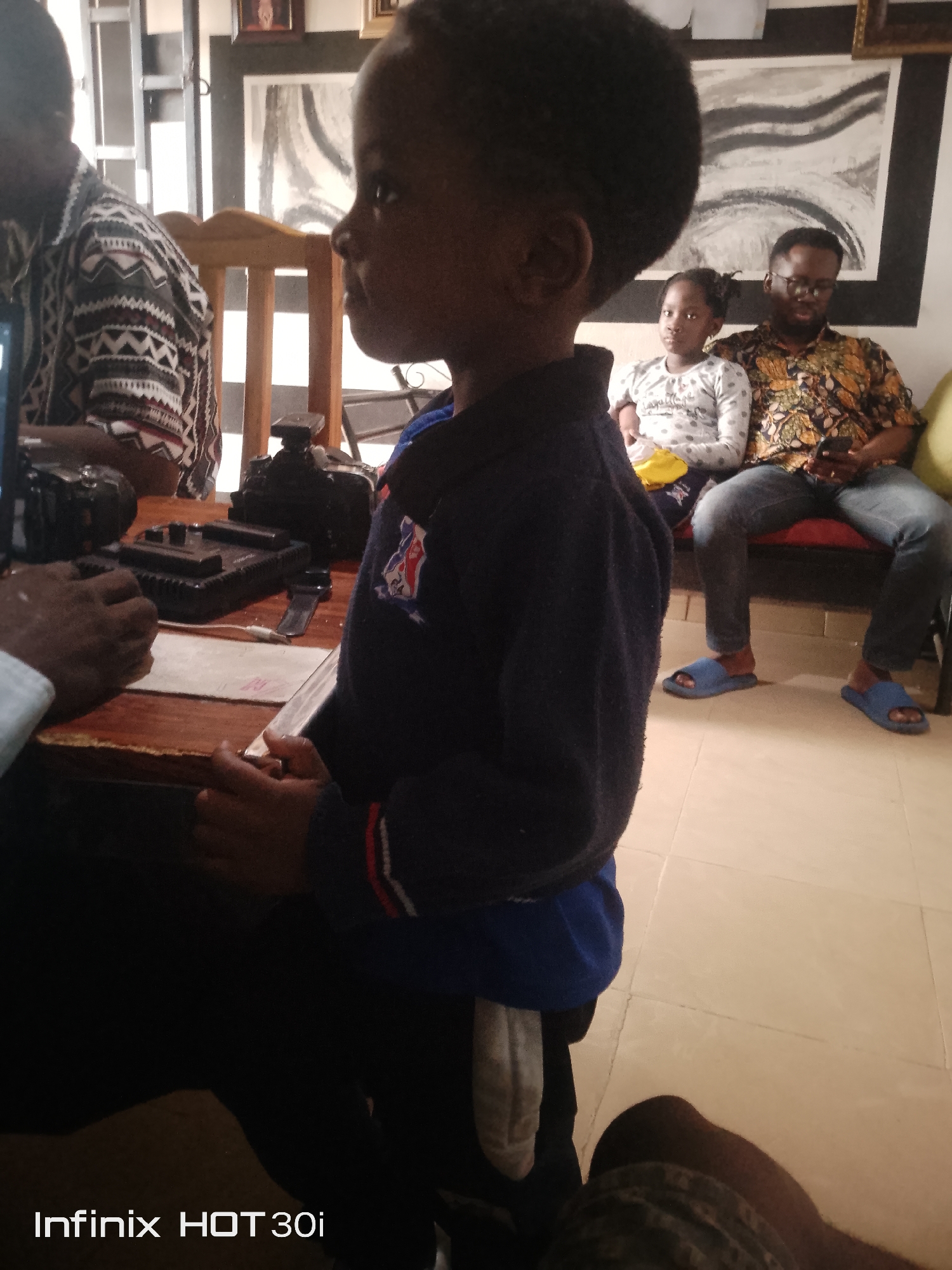 The image shows a group of people gathered around a table, with a young child in the foreground. The child, facing to the left, is wearing a dark blue jacket with a logo on the chest. Behind the child, a woman and a man are seated on a couch. The woman, a young girl, is wearing a white long-sleeved shirt with a polka dot pattern. The man, to her right, is wearing glasses and a colorful patterned shirt. He is holding a mobile phone. On the table in front of them are two cameras and a black electronic device. The setting appears to be indoors, possibly a home, with tiled flooring and artwork on the wall in the background. The lighting suggests it could be daytime. The mood seems relaxed, with the individuals appearing to be engaged in conversation or an activity related to the cameras. The bottom of the image displays the text "Infinix HOT 30i".