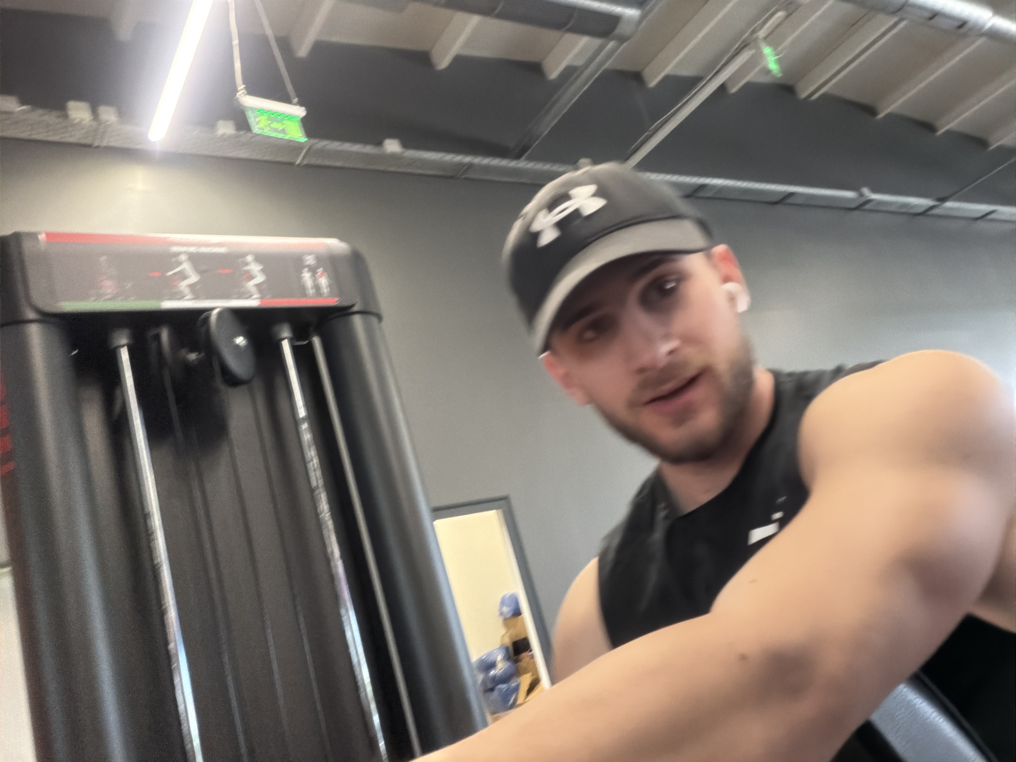 A selfie taken in a gym setting. A man wearing a black Under Armour baseball cap and a black tank top is positioned in the foreground, with his right arm prominently featured. He has short brown hair, a light beard, and is wearing a white wireless earbud in his right ear. He appears to be operating a weight machine, the back of which is visible to the left of the frame. The machine is black with silver detailing and diagrams. In the background, a doorway reveals a glimpse of gym equipment, including blue weights. The location is presumed to be a gym in Plovdiv, Bulgaria. The ceiling is visible and has a grey metal framework with hanging light fixtures.