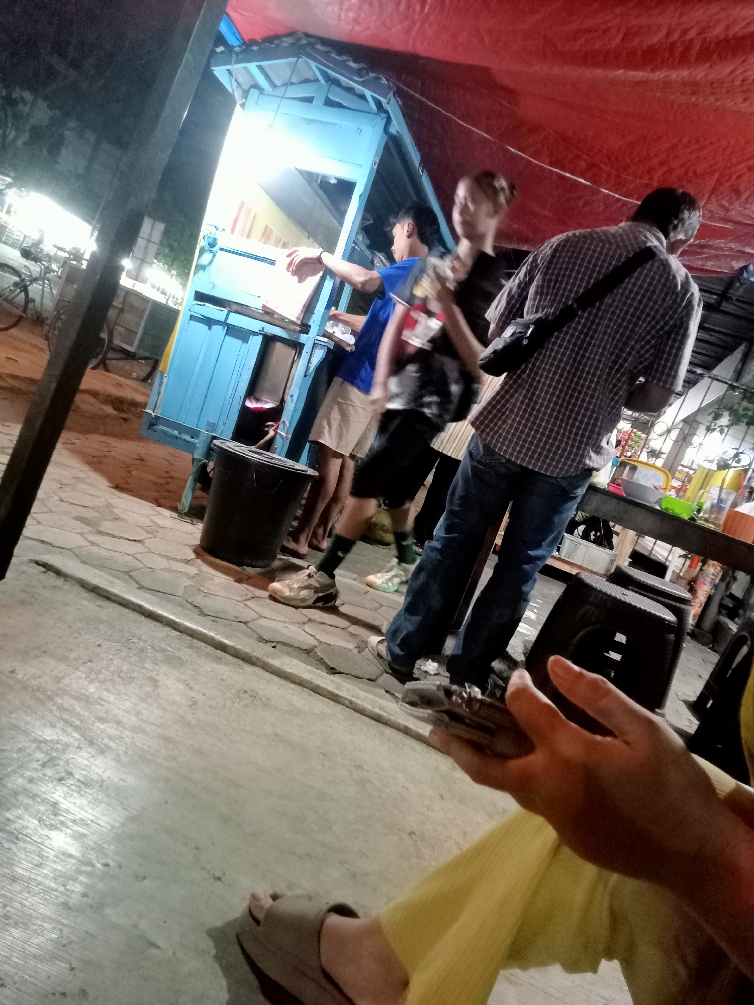 This image depicts a nighttime scene at an outdoor food stall in Mangunsari, Indonesia. The stall is a blue cart with a red tarp covering. Several people are present. In the foreground, a person's hands are visible, holding a phone. Further back, a man in a plaid shirt and jeans is standing. To his left, a person in a blue shirt and shorts is interacting with the stall. Another person, partially obscured, is standing behind them. A black trash can is on the ground near the stall. Bicycles are visible in the background. The ground is paved with hexagonal stones and concrete. The lighting suggests it is evening or night.