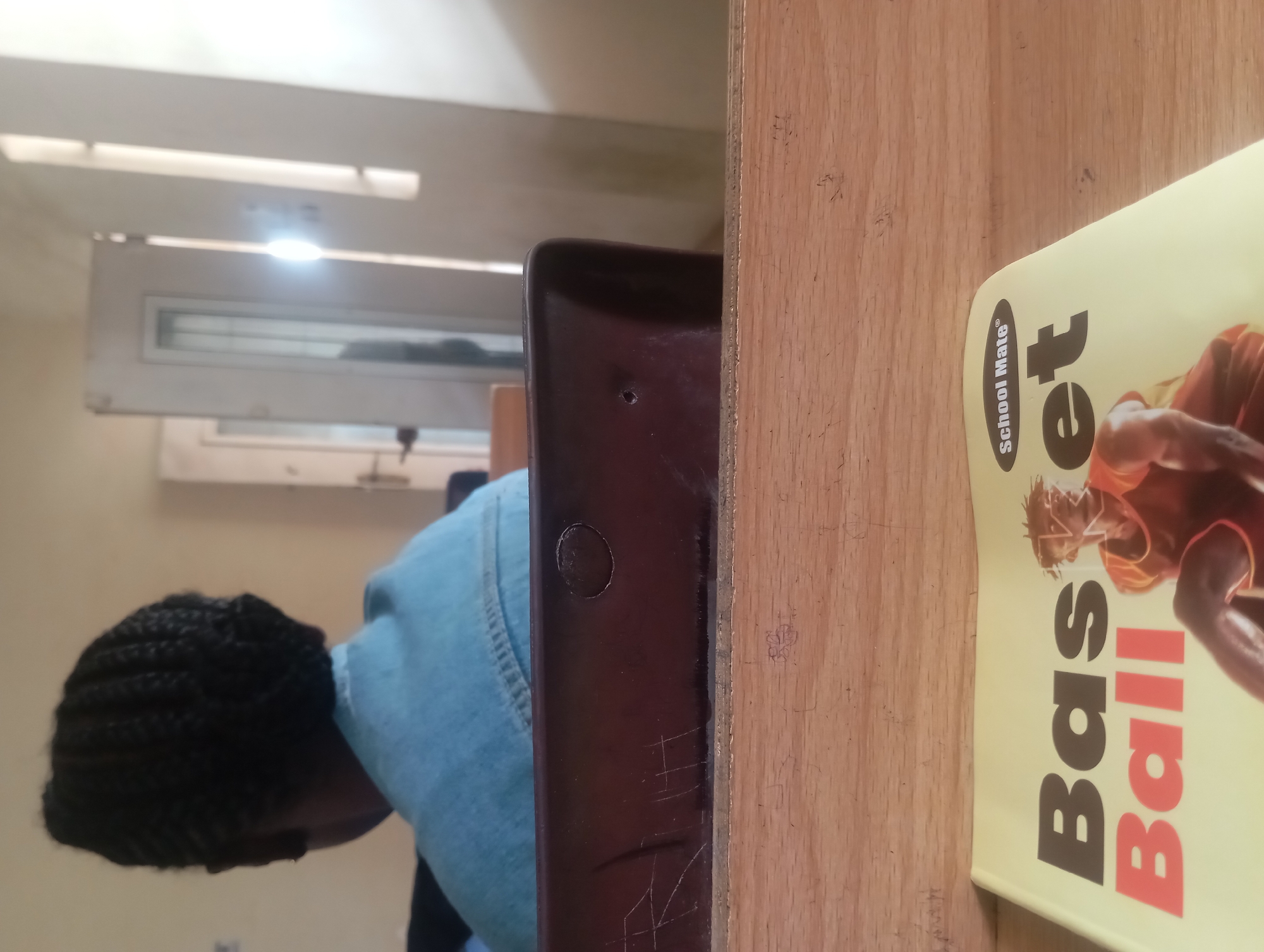 The image, taken from a close-up perspective, shows a book titled "Basketball" resting on a wooden surface. The book is yellow and features the "School Mate" logo. A portion of a person wearing a denim jacket is visible in the background, with their back to the camera. The environment appears to be indoors, with a door and a wall visible behind the person. The lighting suggests daytime.
