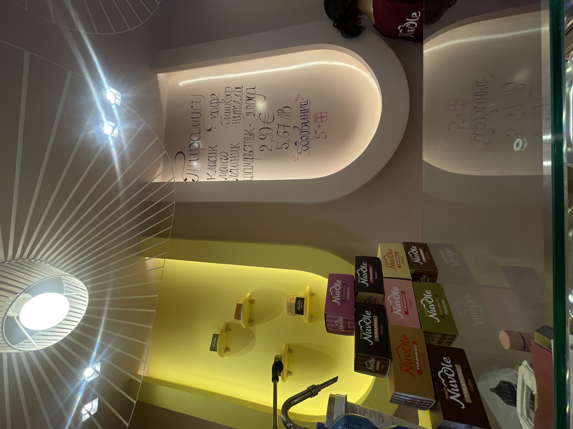 This image shows a display case with "Nuvole" brand tiramisu in various flavors (Classic, Mango, Coffee, etc.) The price list is visible above the product display, written in Bulgarian and English (for Euro pricing). The location is likely a retail shop or cafe in Sofia, Bulgaria, based on the Cyrillic script used on the price list, which includes Bulgarian Lev (MB) pricing, alongside the Euro (€). The environment includes a decorative ceiling and lighting fixtures that complement the shop's interior design.
