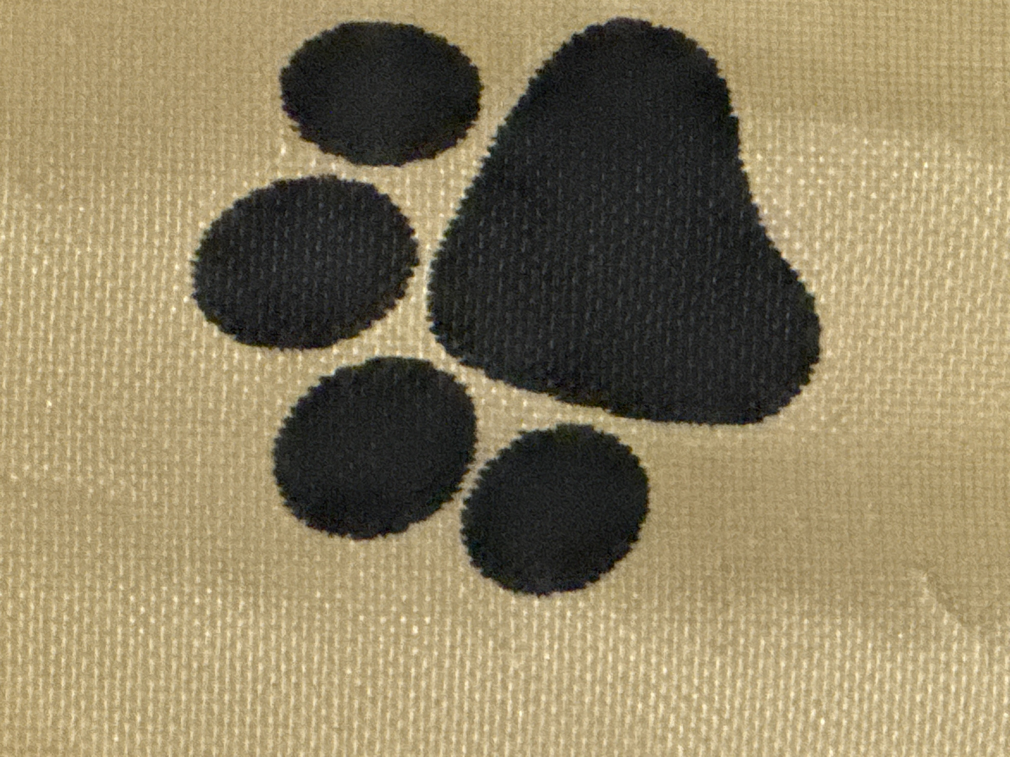 The image shows a close-up of a black paw print emblem on a textured, golden-brown fabric. The fabric appears to be woven, with a visible pattern of horizontal and vertical threads. The paw print is composed of four rounded toe pads arranged above a larger heel pad, characteristic of a dog or cat paw. The edges of the paw print are slightly irregular, suggesting it is embroidered or printed onto the fabric. The lighting is even, and there are no shadows that would indicate a specific environment or time of day. The image provides no information about its location in Sofia, Bulgaria.