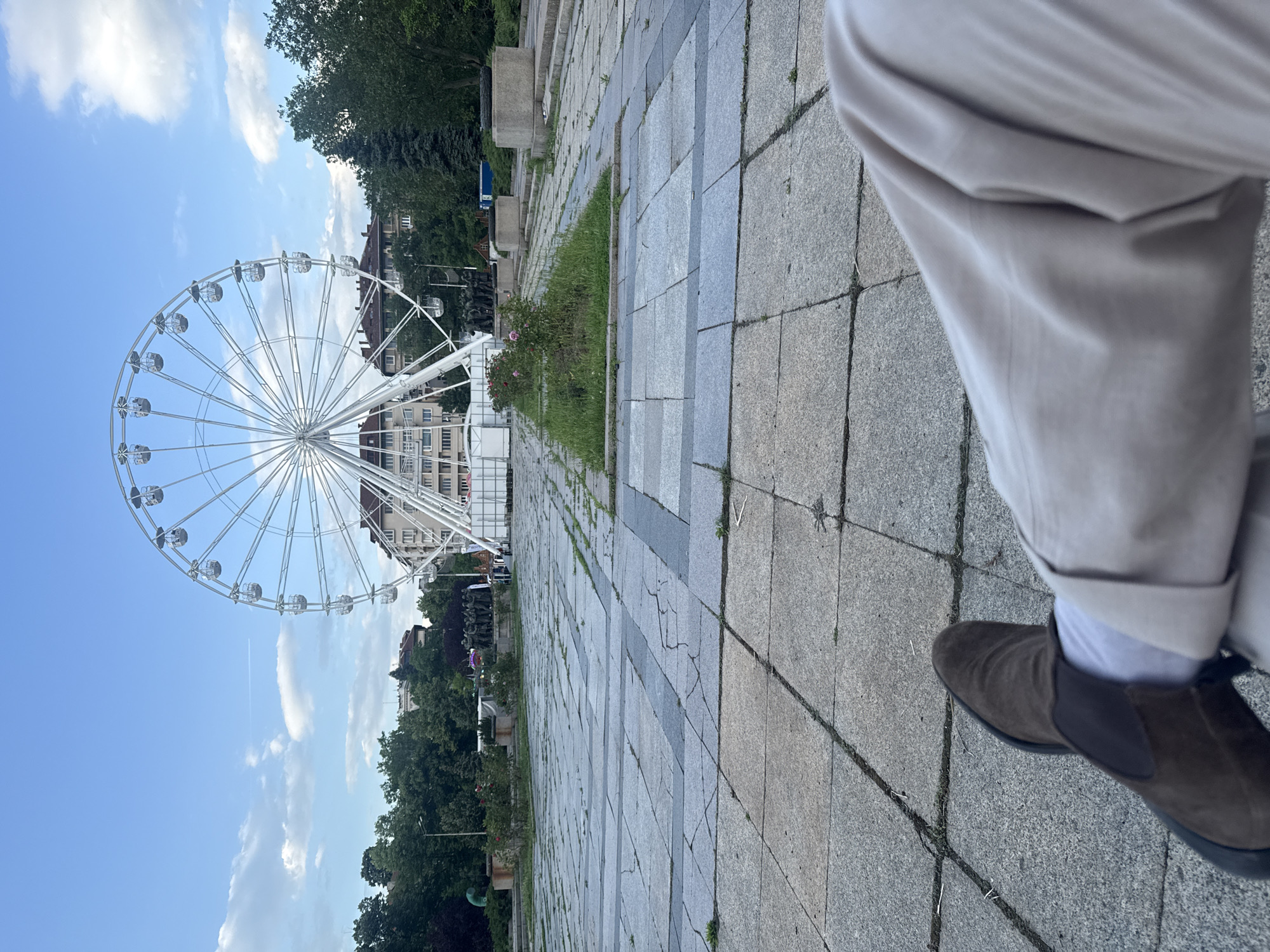 The image captures an outdoor scene, likely a public square or park in Sofia, Bulgaria, on a clear and sunny day with scattered white clouds in a bright blue sky.
In the foreground, on the right side of the frame, the lower part of a person's right leg is visible, wearing light beige or off-white trousers, white socks, and a dark brown suede shoe, possibly a Chelsea boot. The person appears to be standing or walking on a paved surface made of a mosaic of light and dark grey rectangular stone tiles. This paved area stretches from the foreground into the distance, with a narrow strip of green grass and small rose bushes bearing red flowers running alongside it.
On the left side of the image, a large white Ferris wheel dominates the mid-ground. Its intricate spoke structure and numerous transparent gondolas are clearly visible against the sky. Behind and beneath the Ferris wheel, several buildings with multiple stories and dark roofs can be seen, suggesting an urban environment. Green trees are present both behind these buildings and further along the paved area, contributing to a park-like atmosphere. No discernible text is visible in the image.