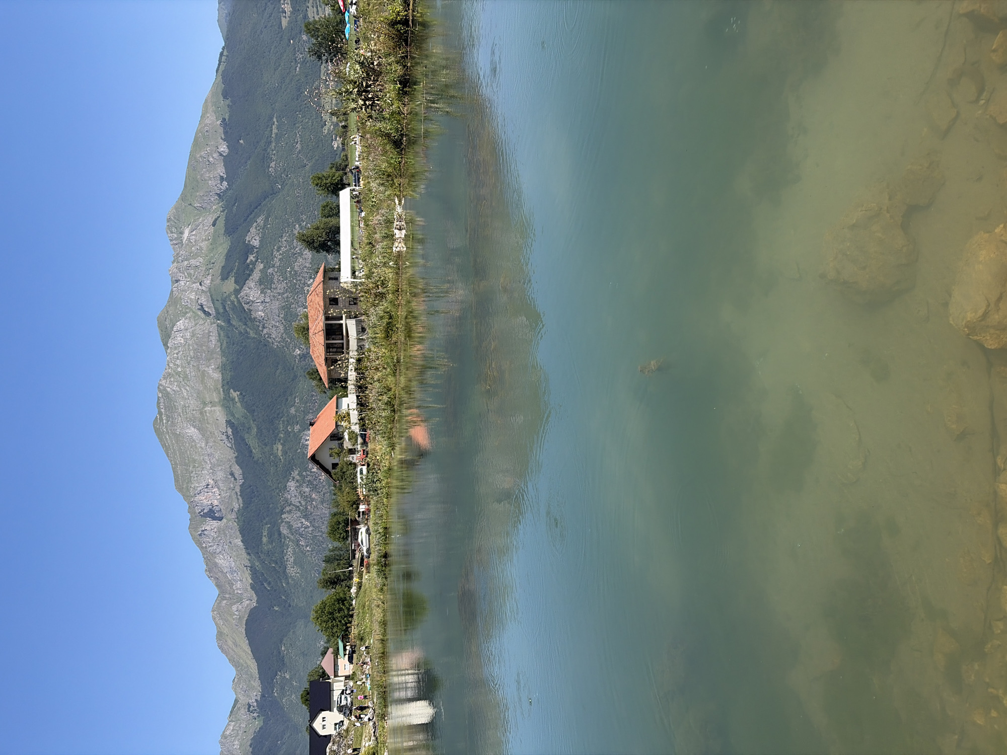 The image shows a serene landscape in Gusinje, Montenegro, with a clear blue sky overhead. A tranquil, turquoise lake dominates the foreground, with its shallow depths revealing submerged rocks and sandy bottom. The water is calm, with subtle ripples indicating slight movement.
On the opposite bank of the lake, a cluster of buildings is nestled against a backdrop of lush green hills that gradually rise to form a majestic, rocky mountain range. The mountains are rugged and imposing, with patches of green vegetation clinging to their slopes.
Several houses with red-tiled roofs are visible along the shoreline, reflecting softly in the lake's surface. Some of these houses appear to be modern residences, while others might be more traditional structures. There are also some people scattered around the area, suggesting a leisurely gathering or a tourist spot. Cars and tents are also visible, further indicating human activity.
The environment appears peaceful and picturesque, with the natural beauty of the mountains and the lake creating a sense of calm and tranquility. The presence of human activity suggests that this is a popular spot for recreation or relaxation.