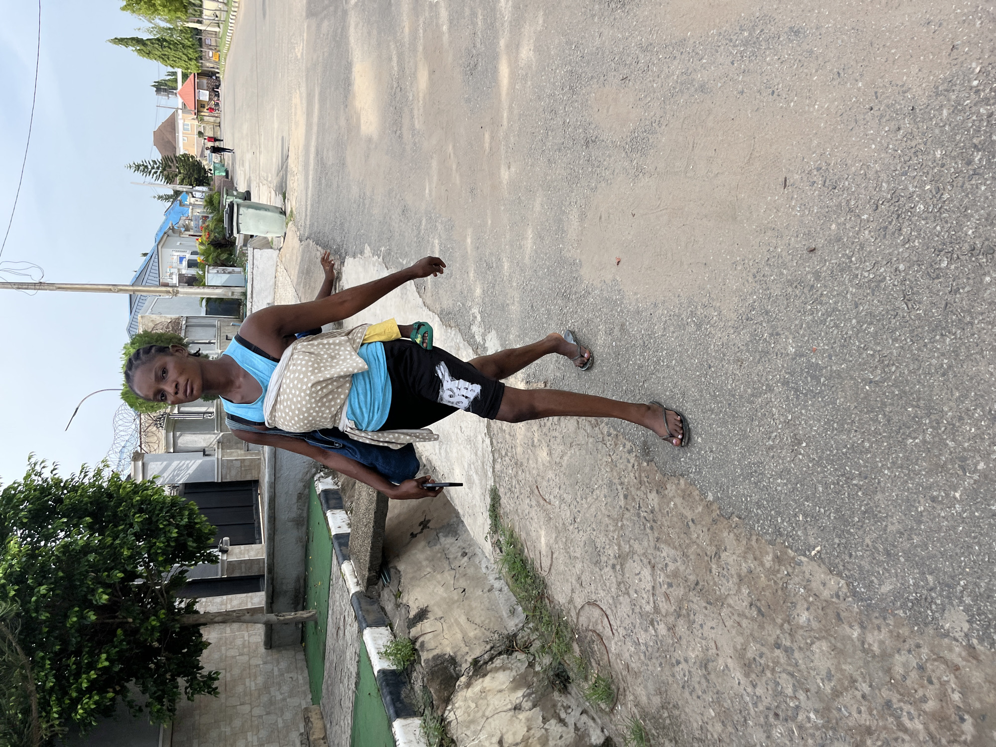 A woman is standing on a street in what appears to be a residential area. She is wearing a blue tank top, black shorts, and sandals. She has a bag slung over her shoulder and is holding a cell phone in her left hand. The street is paved and there are buildings, trees, and a fence with barbed wire in the background. The specific location in Abuja, Nigeria, cannot be determined from the image alone.