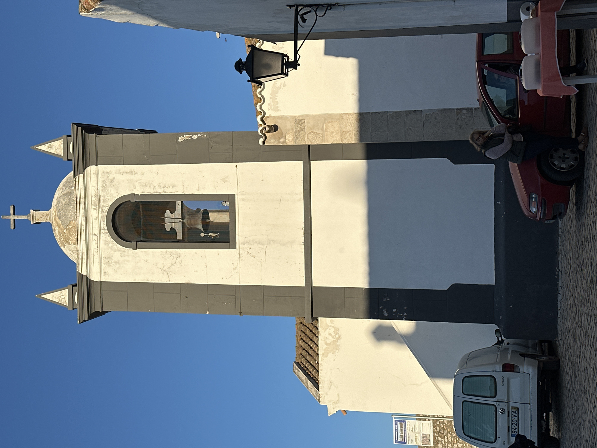 The image shows the bell tower of the Igreja Matriz de Vila Real de Santo António in Vila Real de Santo António, Portugal. The tower is white with dark gray trim and a dome topped with a cross. A bell is visible inside the tower. The tower is situated next to a white building and a street, with a red car and a white van parked on the right side of the image. A woman is near the red car. A black lamppost is attached to the white building. The sky is clear and blue.