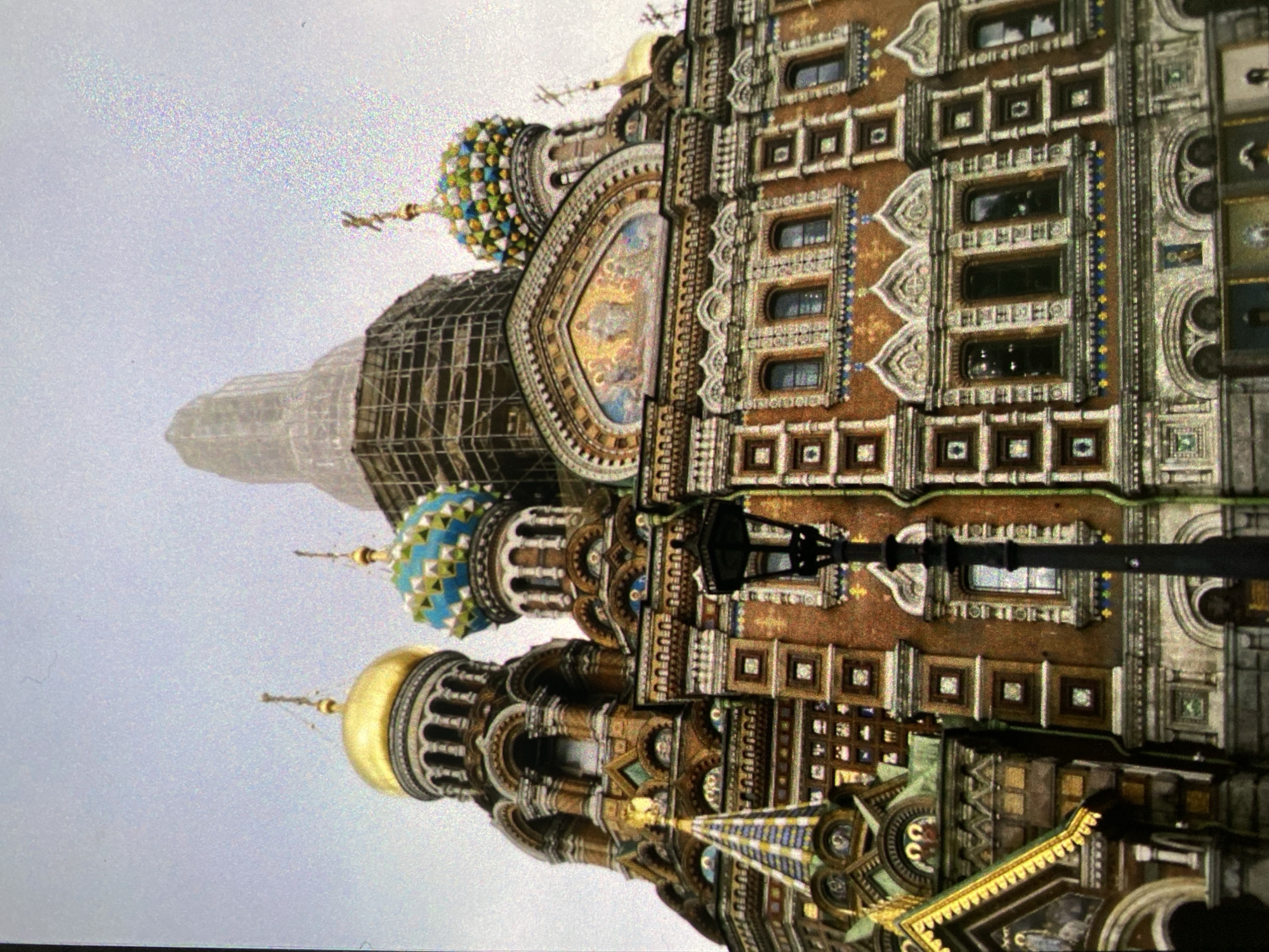 The image depicts the Church of the Savior on Spilled Blood in Saint Petersburg, Russia. The exterior showcases ornate architectural details, including colorful domes, intricate mosaics, and decorative facades. One dome appears to be undergoing restoration with scaffolding partially surrounding it. A lamppost is visible in the foreground. The sky is overcast.