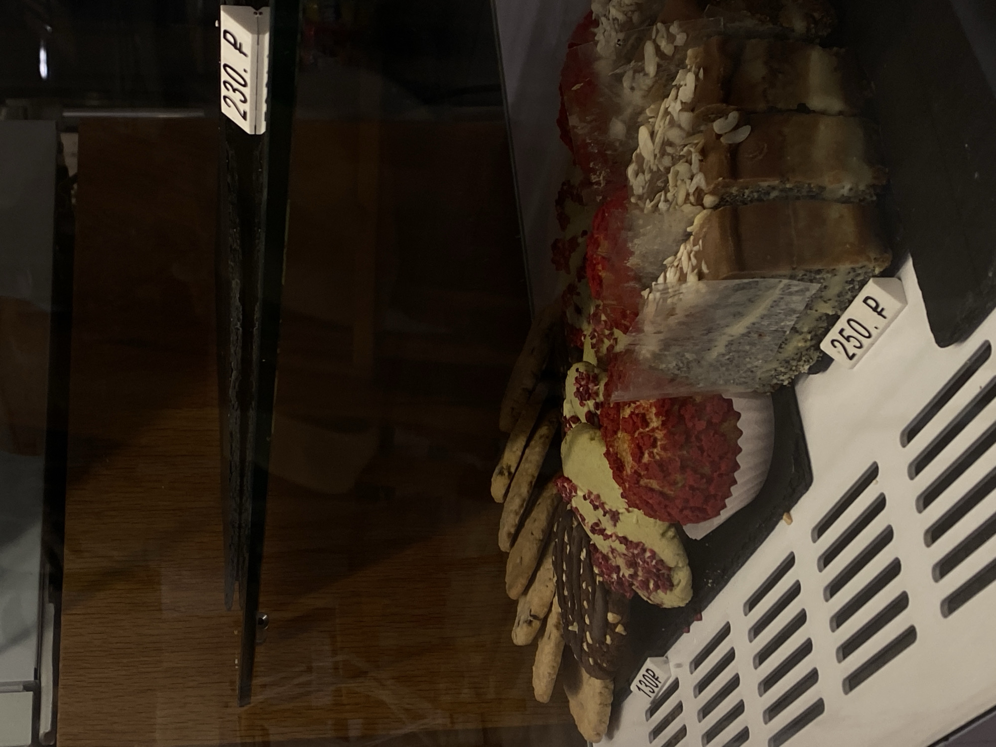 A close-up, low-angle shot showcases a display of baked goods inside a glass-enclosed refrigerated case. The case is illuminated by interior lighting, highlighting the products. In the foreground, a white slatted grate runs horizontally across the bottom of the image. Behind the grate, a black slate slab displays various pastries and cookies.
To the left, a small white price tag reads "130P". A selection of cookies, some topped with chocolate and nuts and others with green icing and red berry pieces, are arranged in rows. Further right, a red, spherical pastry sits in a white paper liner, and next to it are slices of cake topped with almonds and what appears to be poppy seeds. A price tag for the cake reads "250. P". A glass container with a greyish speckled contents is partially visible on the far right.
In the background, a wooden paneled wall forms the back of the display. A glass shelf, slightly smudged, is positioned above the baked goods, with a white price tag reading "230. P" resting on its surface. The overall impression is of a well-stocked bakery display case. The location is identified as Vishnyaki, Russia.