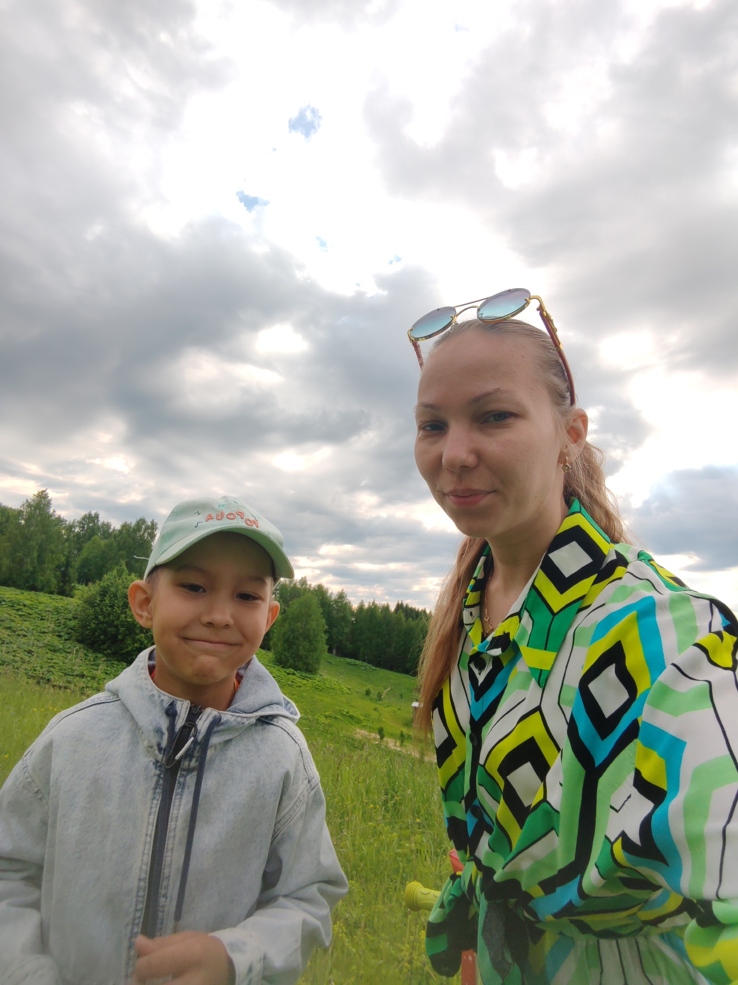 In the city of Chukhlem, Russia, a woman and a young boy are captured in a selfie outdoors. The woman, on the right, has blonde hair pulled back and is wearing a colorful, geometric-patterned shirt. Sunglasses are perched on her head. The boy, on the left, is wearing a light blue baseball cap and a denim jacket. They are standing in a grassy field with trees and a cloudy sky in the background. The boy is smiling at the camera, while the woman is looking slightly to the side. The overall impression is a casual, outdoor portrait.