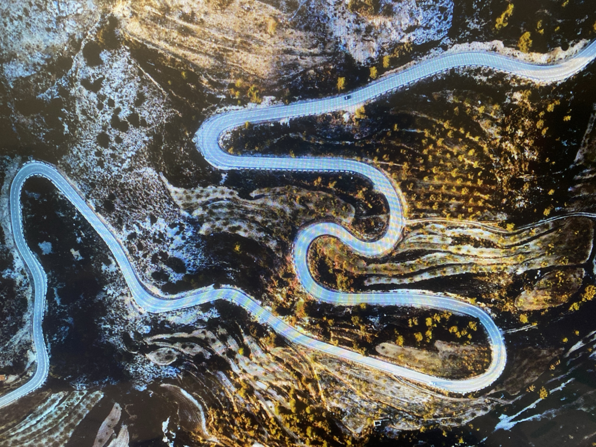 This aerial photograph captures a long, winding road snaking through a rugged, mountainous or hilly landscape. The road features numerous hairpin turns and gentle curves, suggesting it navigates challenging topography. The surrounding terrain is predominantly dark and rocky, with interspersed areas of lighter soil or rock.
Sparse vegetation, appearing as clusters of golden-yellow shrubs or small trees, covers parts of the hillsides, hinting at either an autumn season or a dry, arid environment. The light blue color of the road surface, likely an effect of sunlight and material, contrasts with the darker natural surroundings. A single, dark-colored car is visible driving on one of the road's straighter sections towards the upper-middle of the image.
The scene is illuminated by what appears to be natural daylight, casting subtle shadows that define the contours of the land. The weather seems clear, without visible clouds. The image is a static, high-angle shot, providing a panoramic view of the road's intricate path through the wilderness. There is no visible text.
