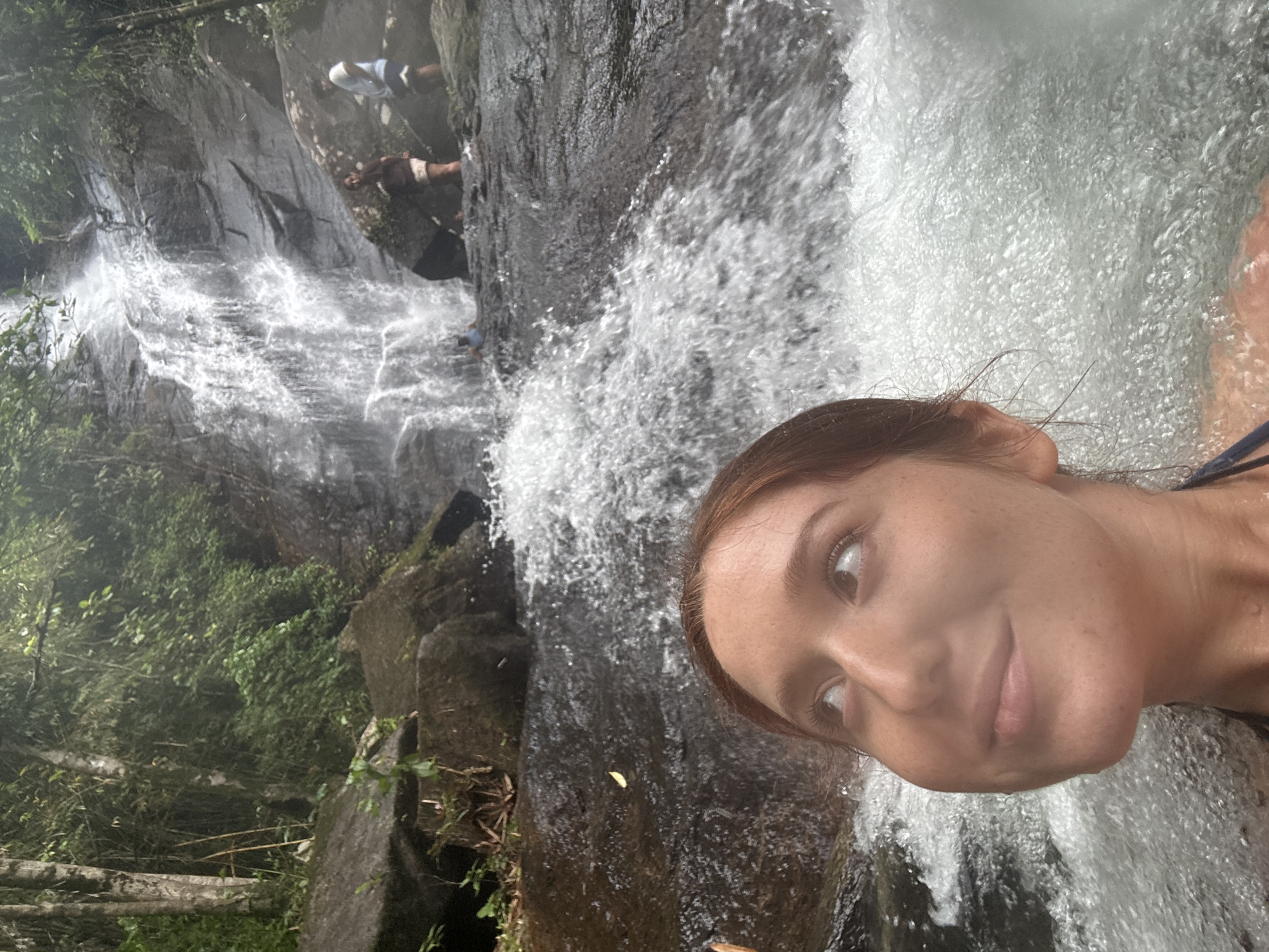A light-skinned woman is positioned in the foreground, with a waterfall cascading behind her. Two indistinct figures are seen standing on a rock face on the left side of the waterfall. Lush vegetation and rock formations flank the waterfall. The setting is possibly in or near Ban Bang Khli, Thailand, due to the tropical characteristics in the background.