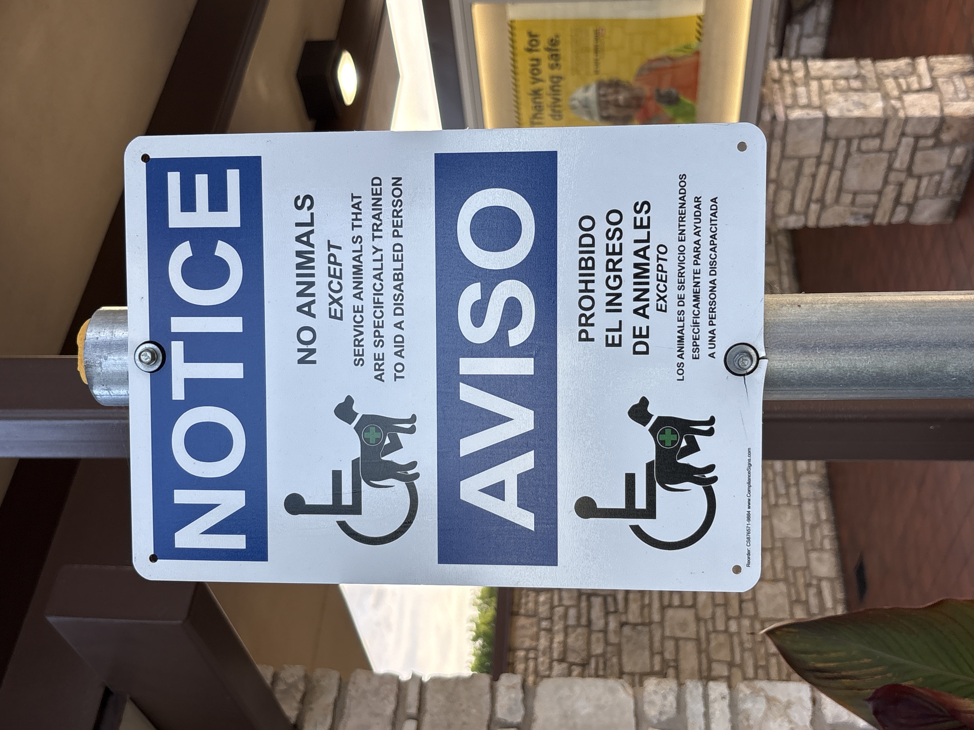 The image shows a bilingual (English and Spanish) sign posted on a metal pole in an outdoor setting, likely a commercial or public area. The sign is white with a blue header that says "NOTICE". Below that, in black text, it states "NO ANIMALS EXCEPT SERVICE ANIMALS THAT ARE SPECIFICALLY TRAINED TO AID A DISABLED PERSON." The sign also features the international symbol of access with a silhouette of a service dog. The Spanish section reads "AVISO" in a blue header, followed by "PROHIBIDO EL INGRESO DE ANIMALES EXCEPTO LOS ANIMALES DE SERVICIO ENTRENADOS ESPECÍFICAMENTE PARA AYUDAR A UNA PERSONA DISCAPACITADA." The background shows a building with stone and dark brown trim, and a glimpse of a yellow sign with text that reads "Thank you for driving safe." The overall scene suggests a location where service animals are permitted, but other animals are not. The specific location is a parking lot in Sonora, United States.