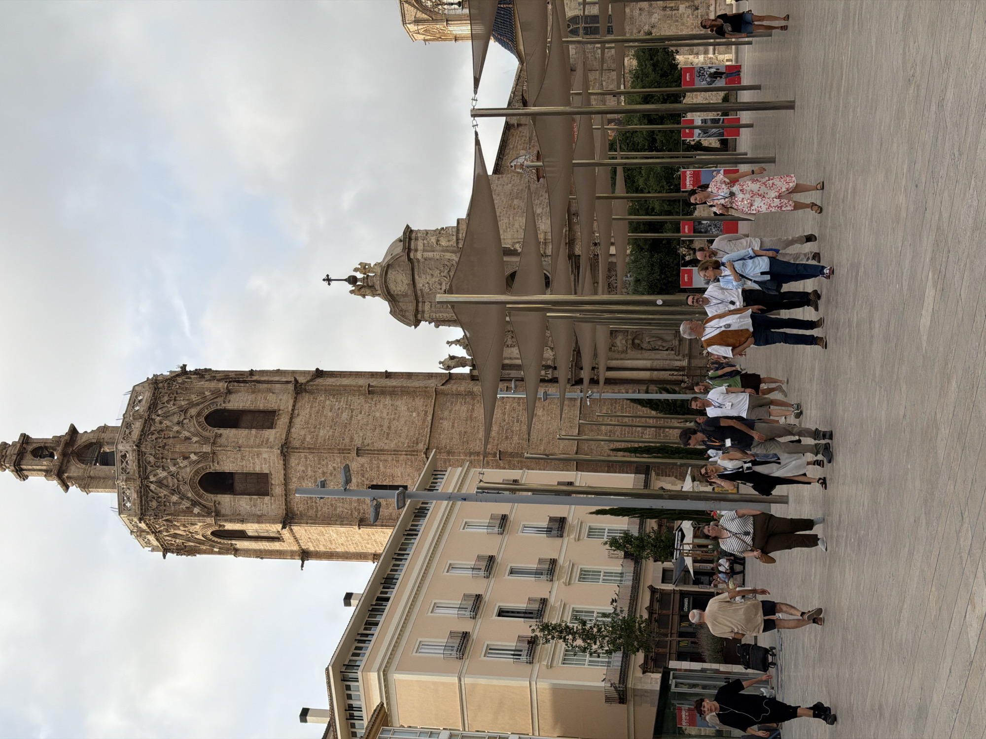 An outdoor scene in a European city square features a tall, ancient stone bell tower and a large, ornate building with multiple arched windows and decorative stonework. In the foreground, a paved plaza is populated by several groups of people walking. To the left, a light-peach colored building with many windows and balconies is visible. Several trees are interspersed throughout the scene. The sky is overcast.