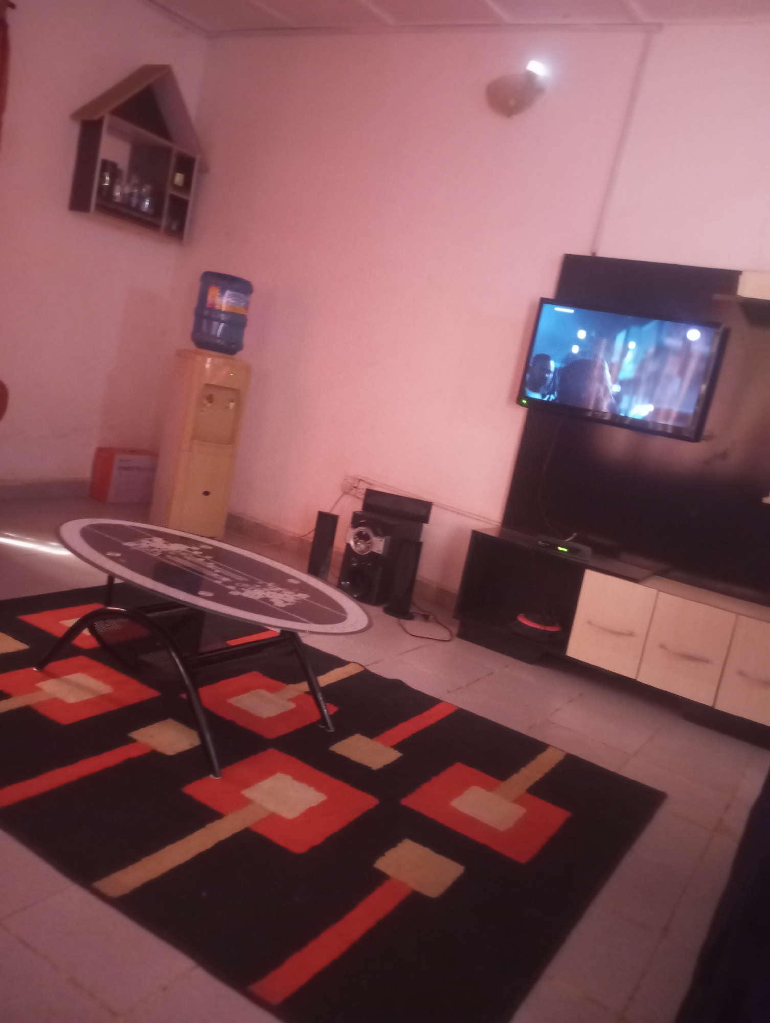 The image depicts an indoor scene, likely a living room, in Yelwa, Nigeria. The room is decorated with a black rug featuring a geometric pattern of red and beige squares and rectangles. An oval-shaped table with a dark top and a black metal base sits on the rug. To the left of the table is a tall, light-colored water dispenser with a blue water jug on top. A small, orange box is visible behind the water dispenser.
In the background, a television is mounted on the wall, displaying an image. Below the television is a black entertainment unit with several compartments, two of which are open and one is closed with a white drawer front. To the left of the entertainment unit, a black speaker is placed on the floor. Above the water dispenser, a triangular shelf unit is mounted on the wall, containing several small items. The walls of the room are painted a pale pink.