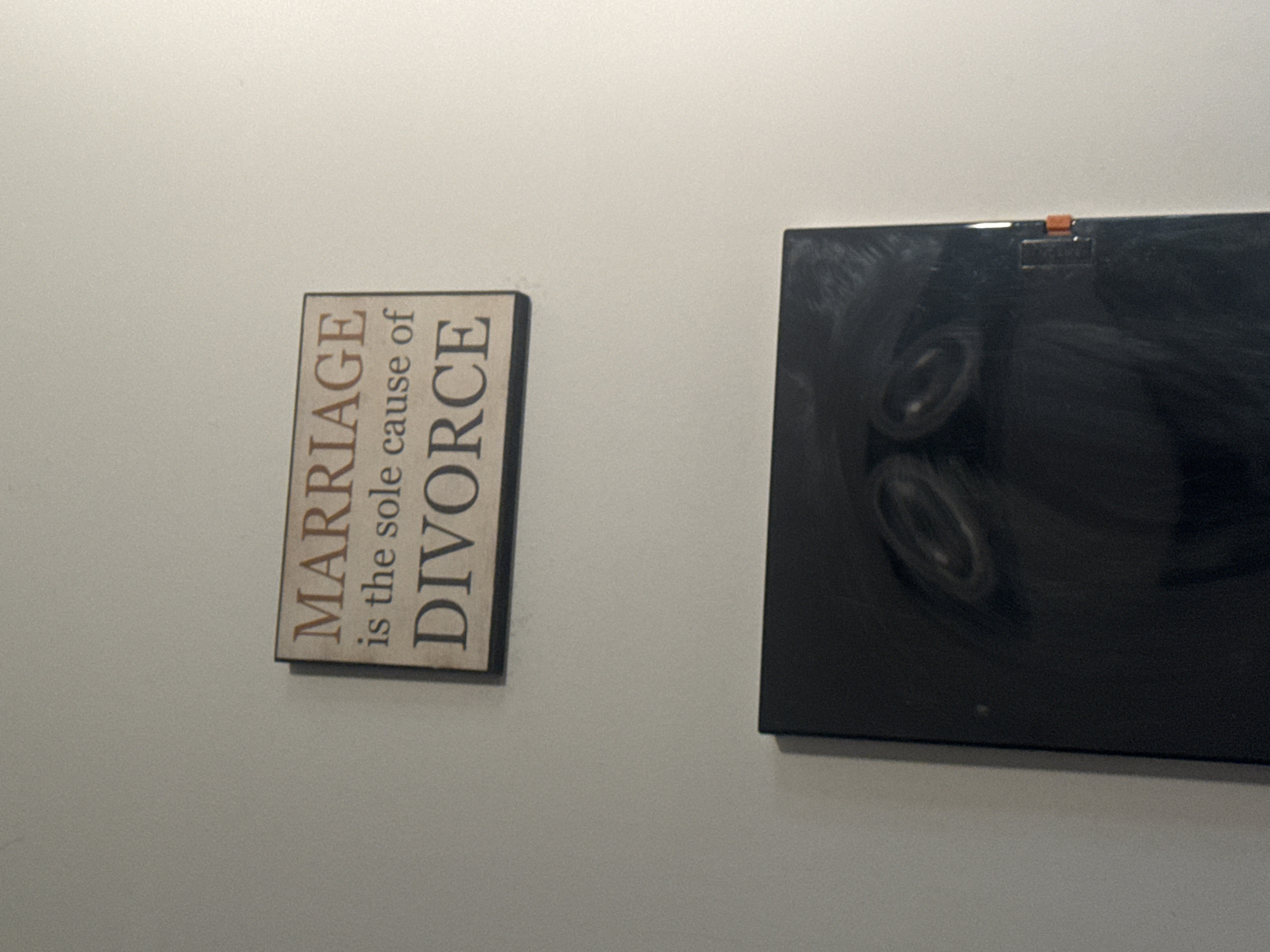 A wooden sign with the words "MARRIAGE is the sole cause of DIVORCE" is mounted on a white wall. Below the sign is a black, rectangular object with two circular indentations on its surface. An orange tab is visible on the right side of the object.