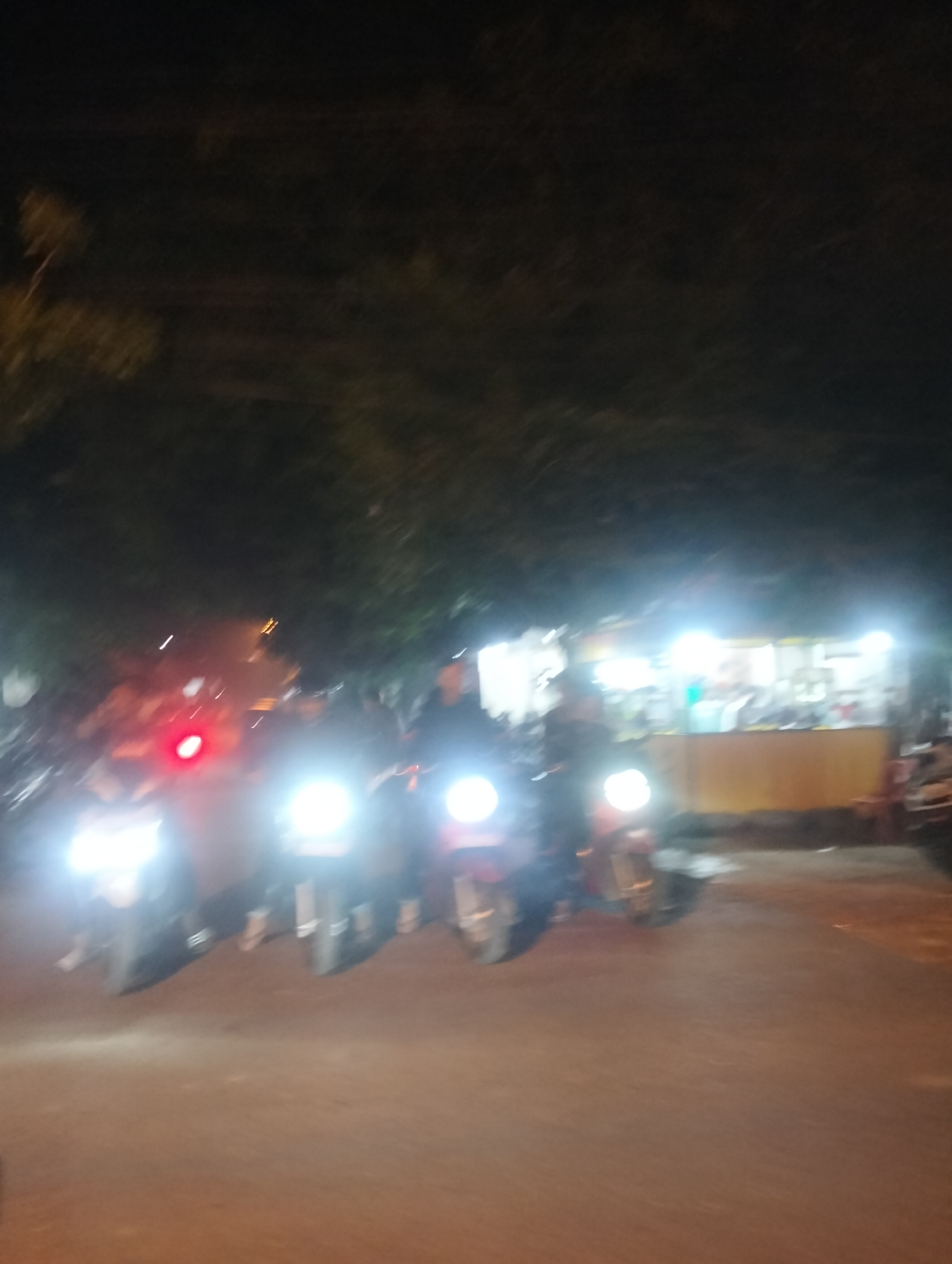 The image depicts a nighttime scene on a street in Lhokseumawe, Indonesia, likely in the vicinity of a food stall or small shop. Several motorcycles are parked or moving, their headlights illuminating the foreground. In the background, bright lights emanate from what appears to be a roadside establishment, possibly selling food or drinks. The atmosphere is dark, with trees visible in the upper portion of the frame, suggesting an outdoor urban setting. The focus is on the vehicles and the illuminated storefront, with blurry figures of people present but not clearly discernible. The ground is paved, indicating a street or walkway. The overall impression is of a casual, late-night gathering or transit point.