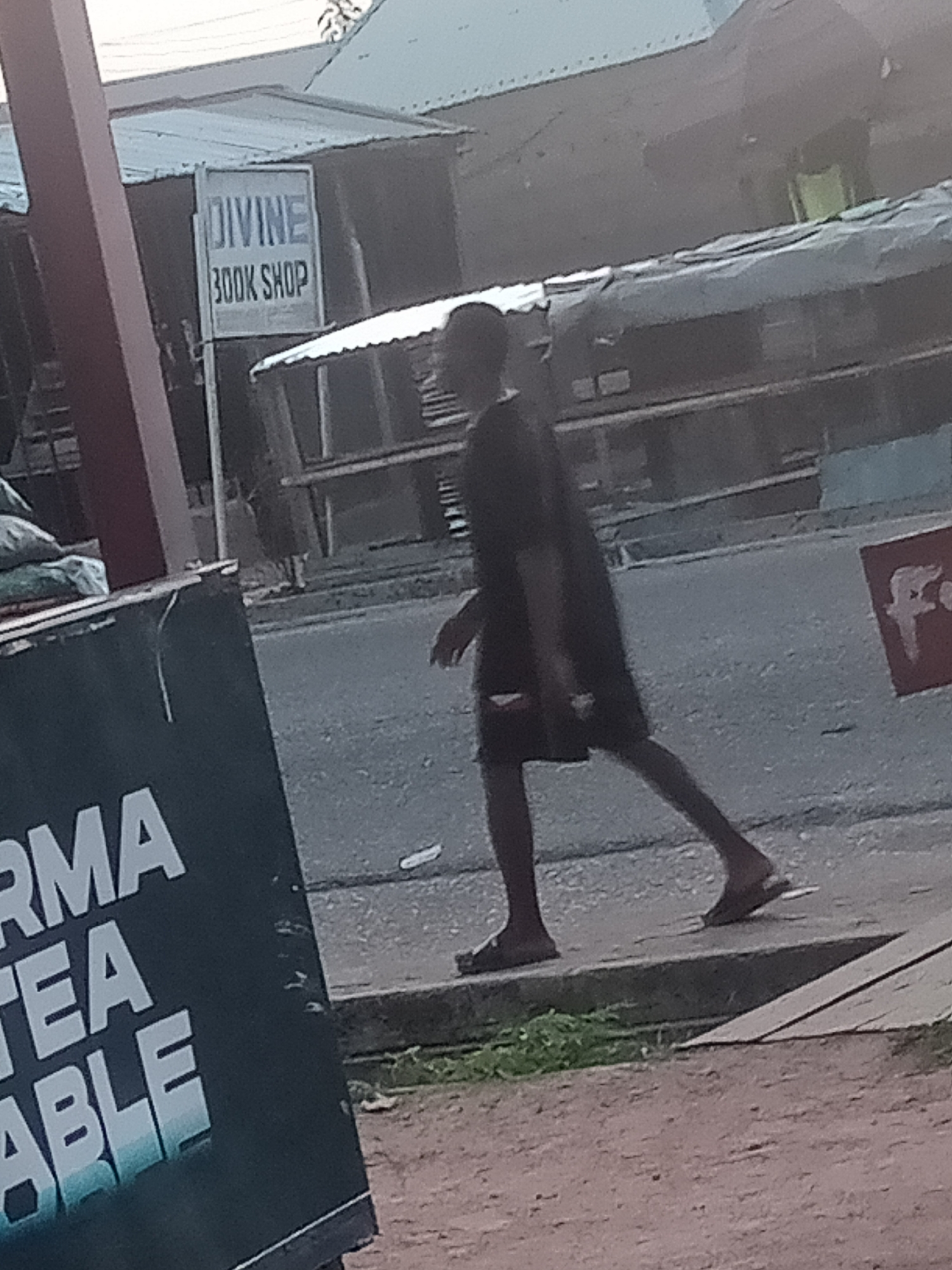 A person walks on a paved surface next to a road. The person is wearing dark clothing and sandals. In the background, there is a sign that reads "DIVINE BOOK SHOP". To the left of the scene, a dark sign with white text is partially visible. The environment appears to be an urban area, likely in Jalingo, Nigeria, with buildings and street infrastructure visible. The lighting suggests it is daytime.