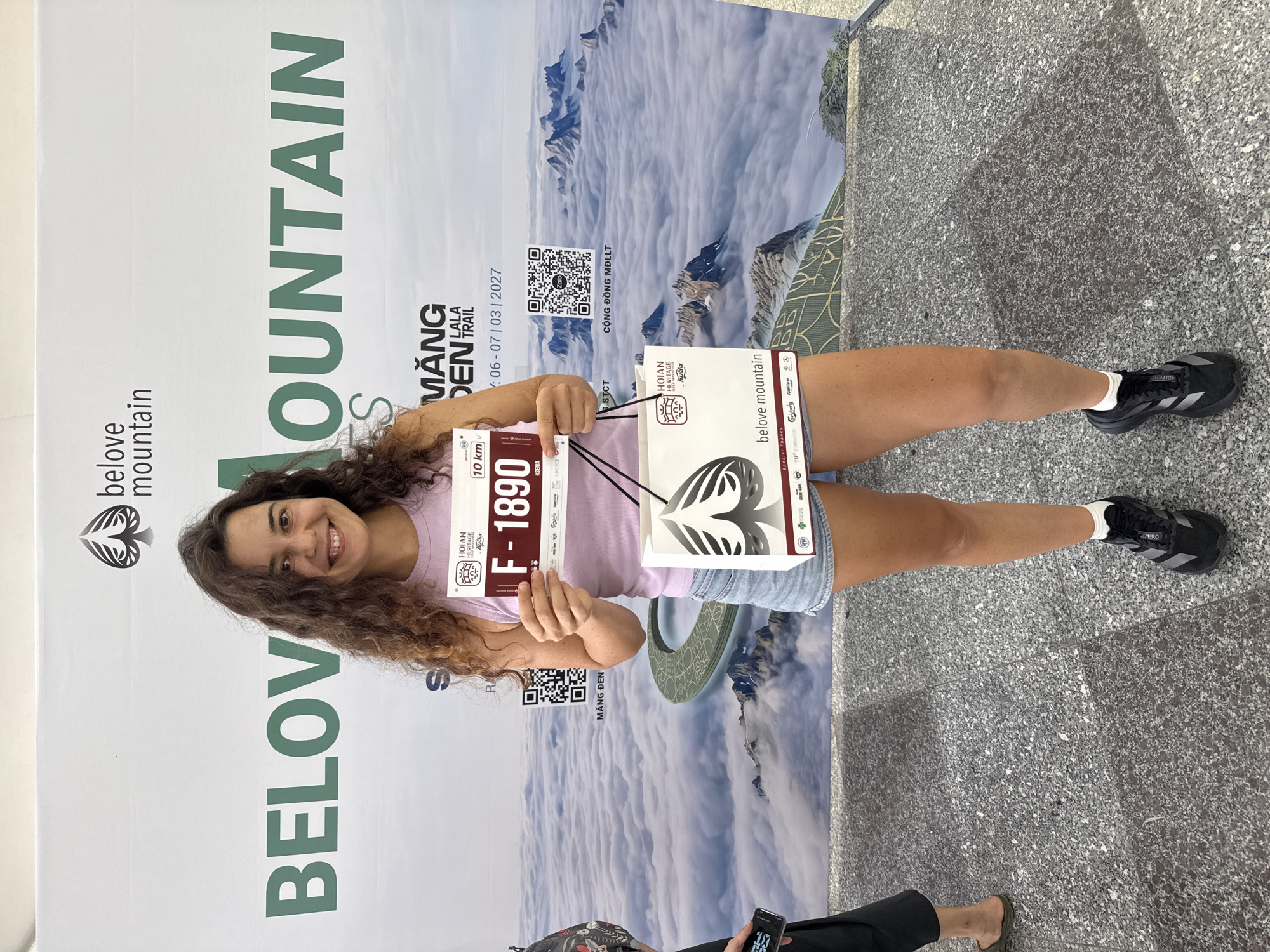 In Hoi An, Vietnam, a young Caucasian woman with curly brown hair stands in front of a large banner displaying "BELOVE MOUNTAIN." She is wearing a purple tank top and denim shorts, holding a running bib with the number "F-1890" and a shopping bag featuring the "belove mountain" logo. The banner features a mountainous landscape covered in clouds and the text "MANG DEN LALA TRAIL" and dates "06-07 | 03 | 2027." She is positioned on a grey granite tiled floor, with other attendees visible in the background.
