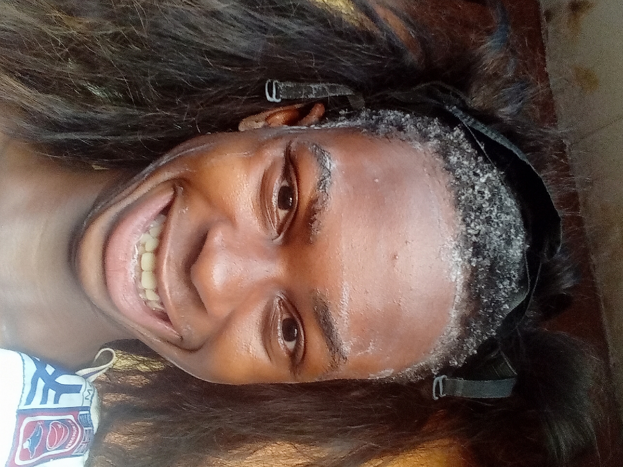 A person with dark skin and long, dark hair smiles broadly at the camera. They are lying on their back. The person's forehead appears to be covered in a white, granular substance. A black headband with adjustable straps is visible. The image may have been taken indoors, given the lighting and apparent lack of background details. There is a towel with a design and writing on it visible in the lower left corner. No context exists in the image to establish the location in Nkpor, Nigeria.
