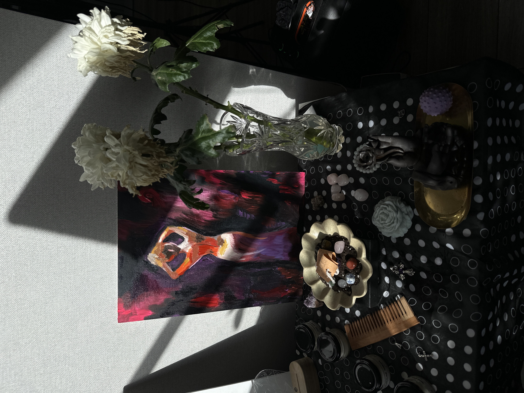 This overhead shot presents an indoor still life arrangement, dramatically lit by direct sunlight casting strong shadows. The scene is set across two surfaces: a light grey, textured surface on the left and a black tablecloth adorned with white polka dots (some solid, some hollow circles) on the right.
On the left, two white, somewhat wilted chrysanthemums with green leaves are displayed in a clear, cut crystal vase, which also contains water and possibly a lime slice. Below the vase, a painting depicts a stylized female figure in a reclining or stretching pose, rendered in warm red and orange hues against a dark background of purple and red brushstrokes. Diagonal bands of sunlight illuminate parts of the grey surface and the flowers.
On the right side of the image, various items are arranged on the polka-dotted tablecloth. A golden rectangular tray holds a dark, hand-shaped sculpture, possibly an incense holder, and a small, purple spiky ball. Nearby, a light-colored, ruffled ceramic bowl contains an assortment of small crystals and polished stones, including pink (likely rose quartz) and purple (likely amethyst) varieties, along with a small paper where the word "HEART" is visible. Several loose, polished pink stones are scattered on the tablecloth. A white, rose-shaped candle or decorative item, a wooden comb, and multiple small glass jars with dark lids are also present. Text on one jar clearly reads "BALM" and "BLACK", with "BALM" visible on another. Two small, silver-colored cross ornaments complete the arrangement. In the upper right background, electronic equipment is partially visible in shadow.
The strong, directional sunlight suggests the time of day is either morning or late afternoon. The arrangement conveys a contemplative or personal space, combining natural elements with artistic and spiritual objects. The location context is Murino, Russia.